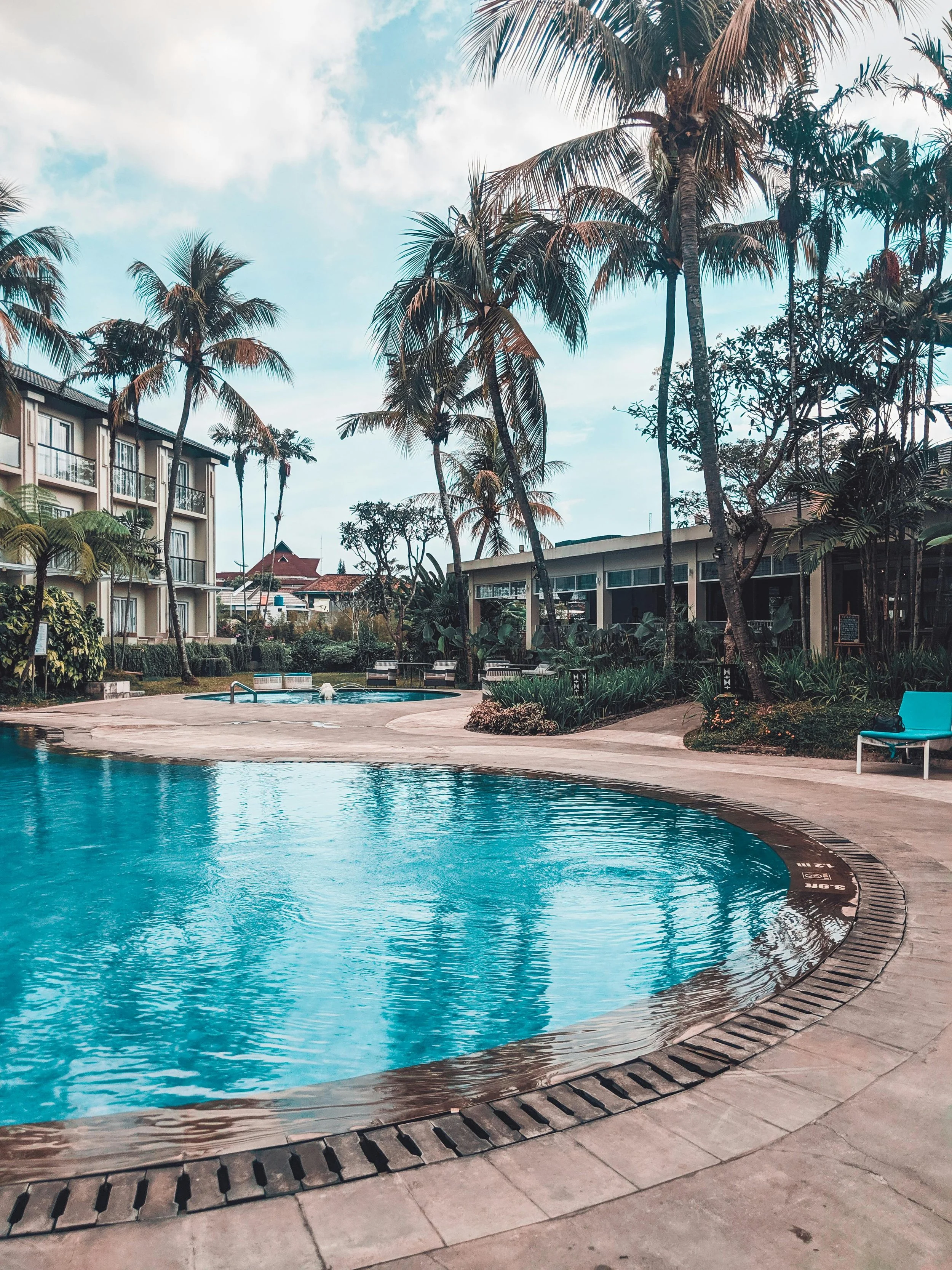 A swimming pool surrounded by palm trees, lounge chairs, and a hotel or resort building, under a partly cloudy sky.