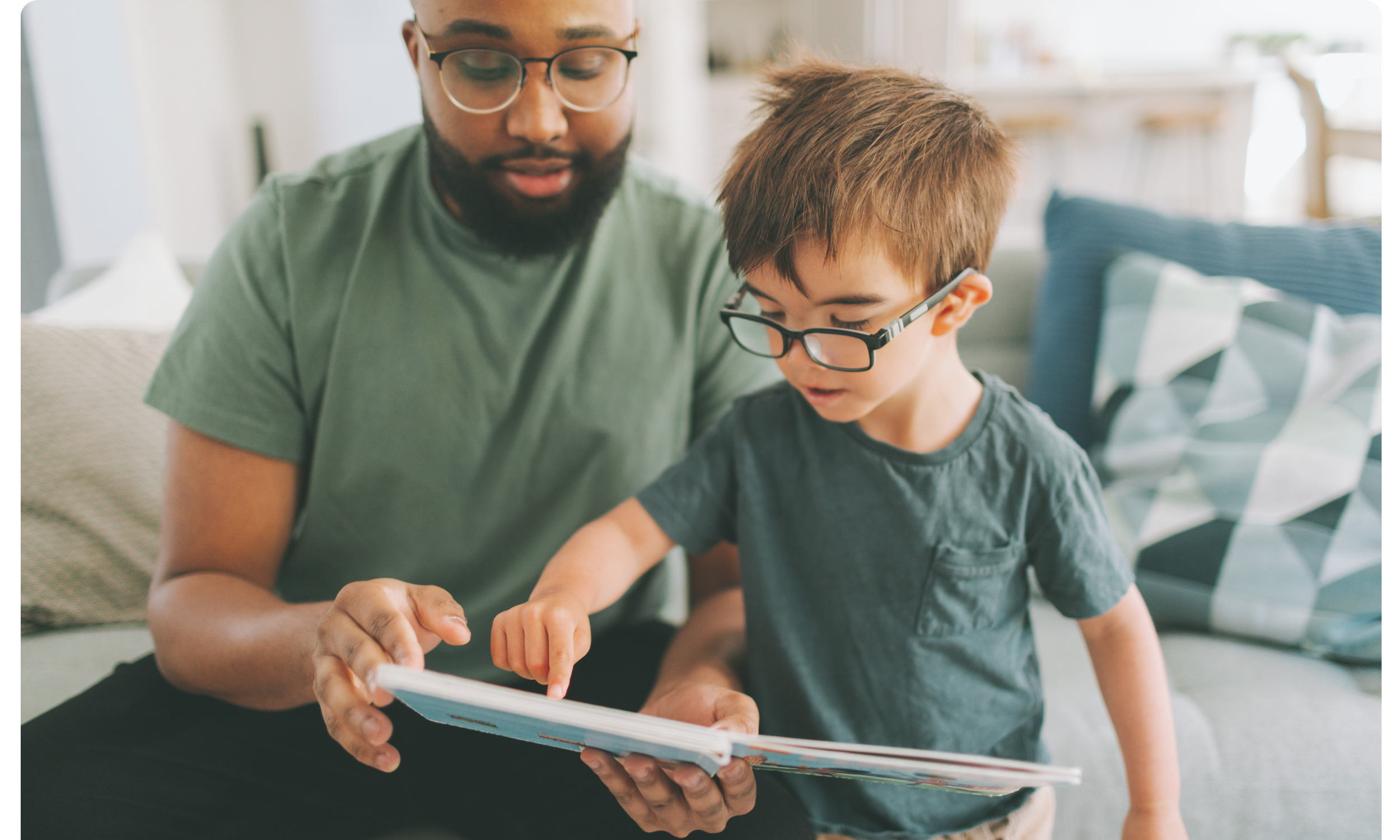A man and young boy with glasses sitting on a sofa, looking at a book together in a bright living room.