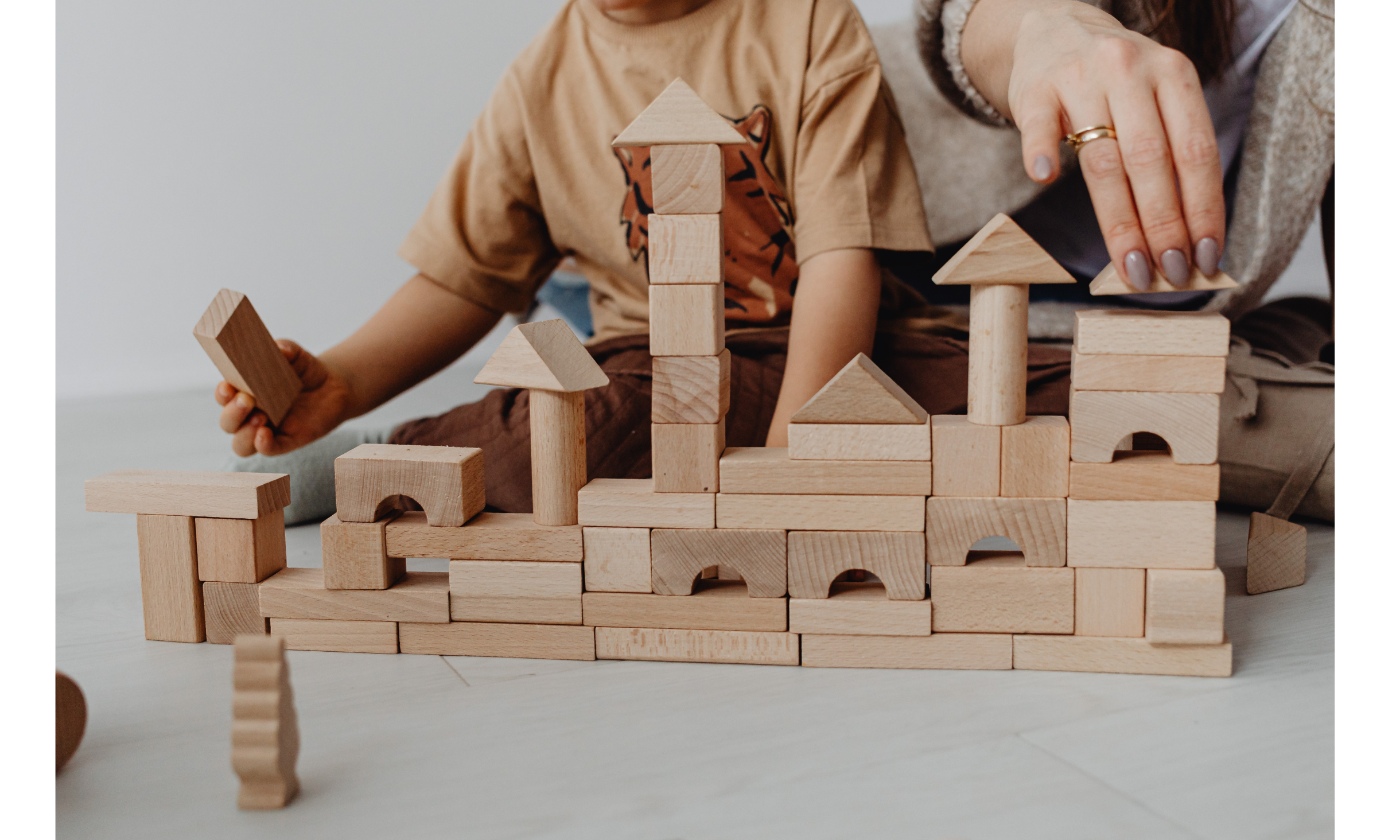 Child and adult building a block tower with wooden blocks on a light-colored floor.