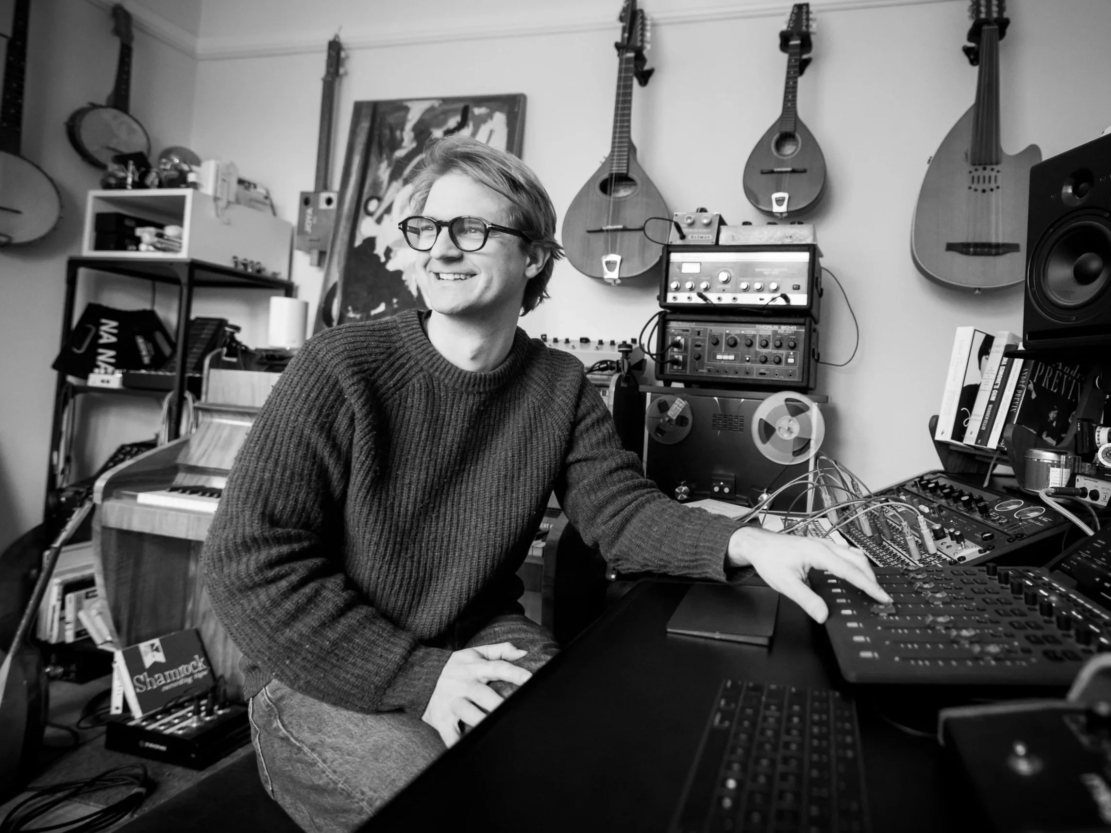 Alex Baranowski in his studio smiling in a music studio surrounded by guitars, recording equipment, and books.