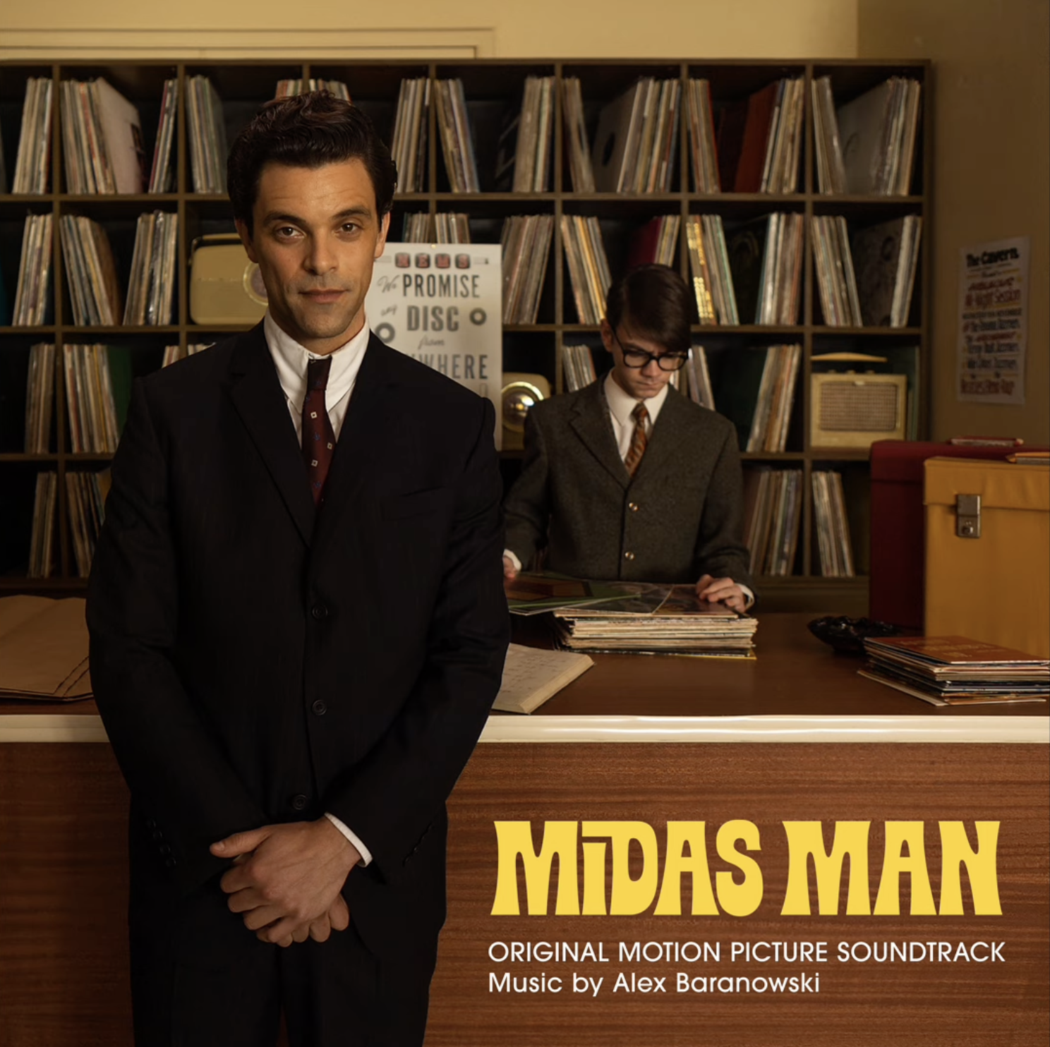 Poster for the movie 'Midas Man' featuring a man in a black suit and tie standing in front of a library counter, with a young man in a blazer and glasses browsing records in the background.