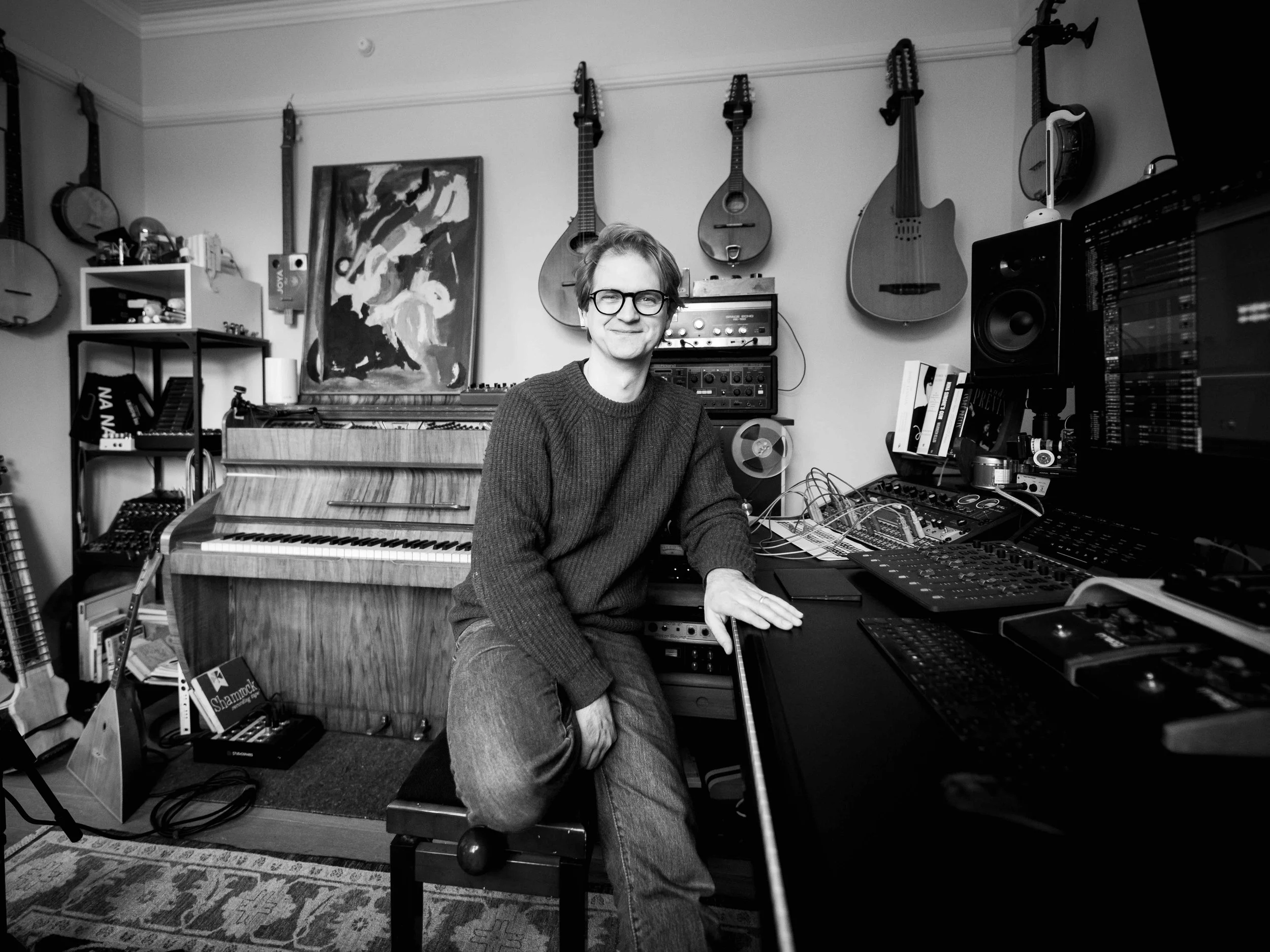 Alex Baranowski sitting in his music studio surrounded by guitars, a piano, and audio equipment.