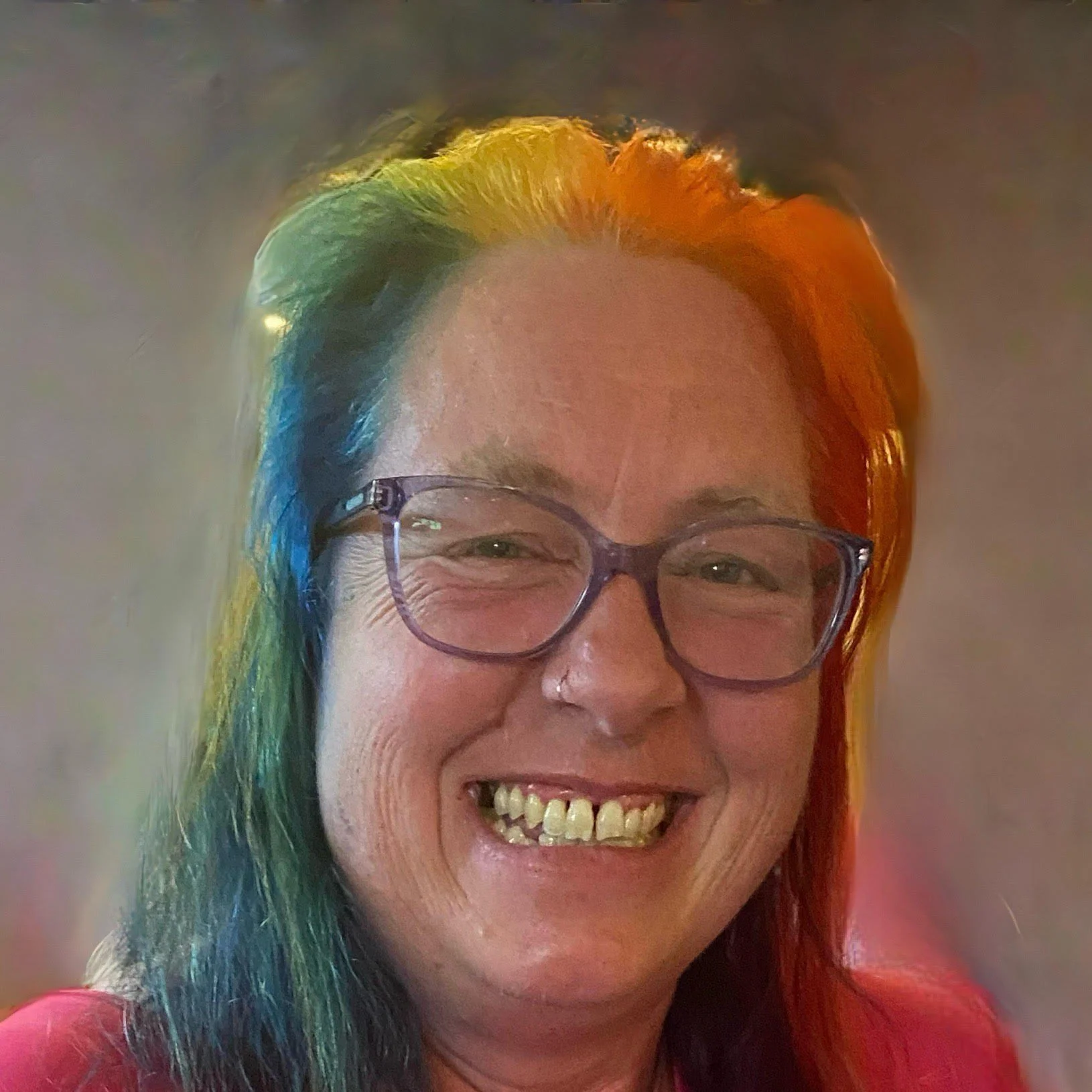 A woman with rainbow-colored hair, wearing glasses, smiling and showing her teeth.