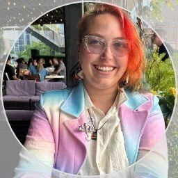 A young woman with red hair and glasses smiling at the camera, sitting in a cafe or restaurant with people and greenery in the background.