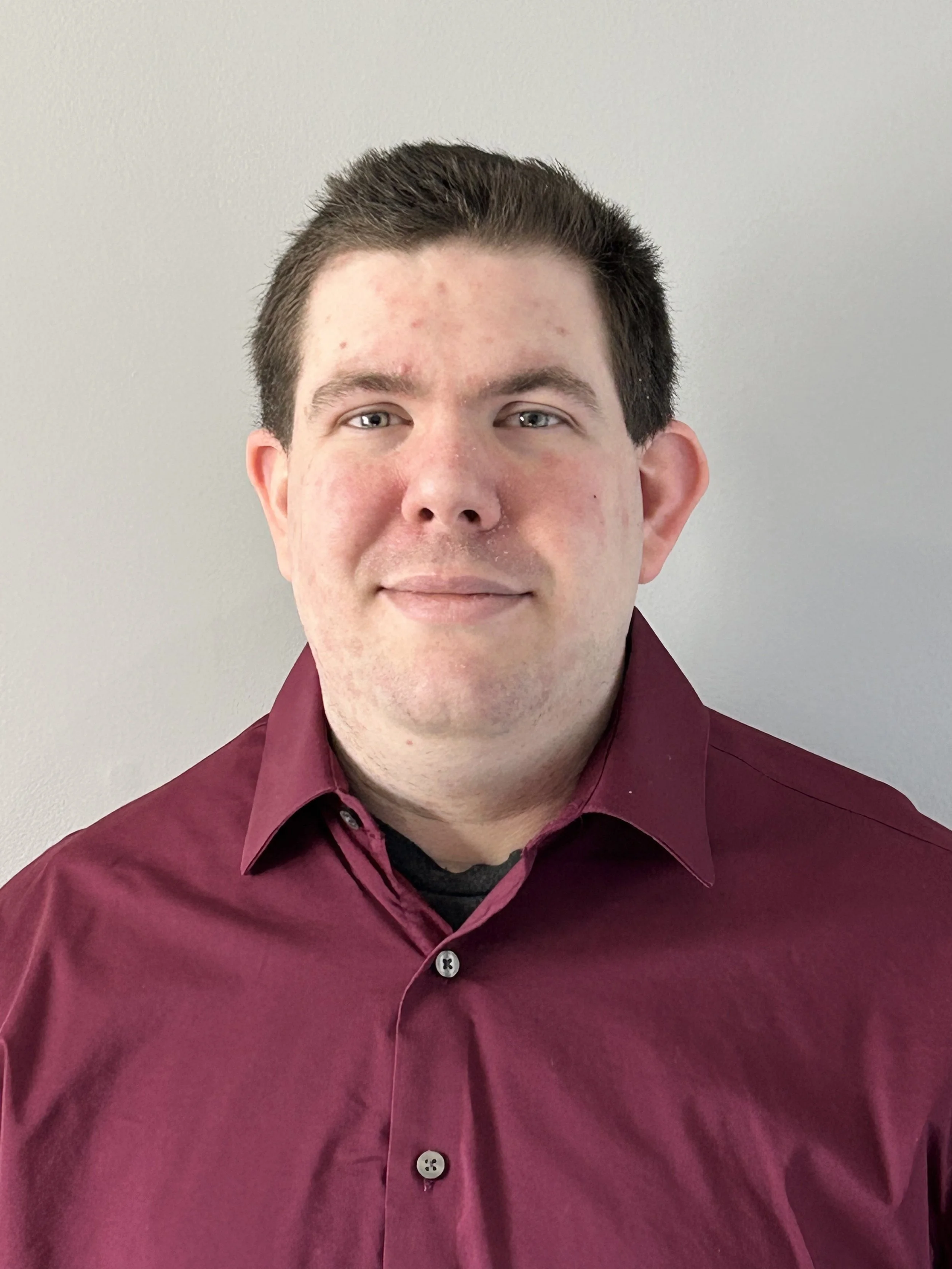 A man with dark hair, wearing a maroon button-up shirt, standing against a plain light gray wall.