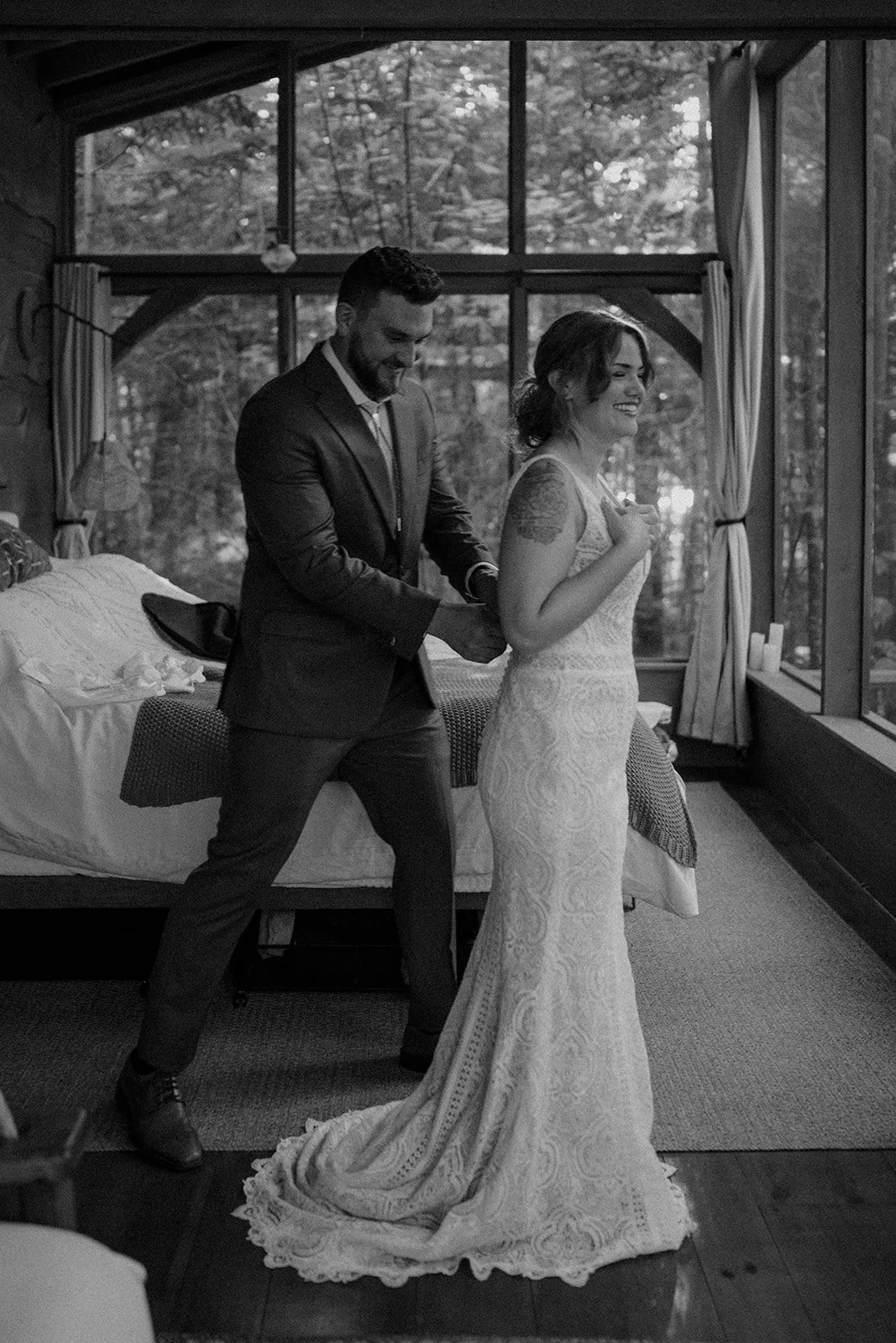 bride and groom getting ready together in front of a large lakeside cabin window