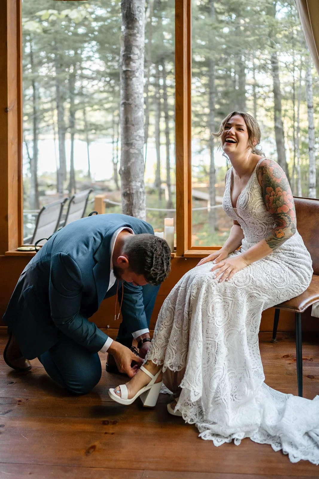 bride and groom getting ready together in front of a large lakeside cabin window