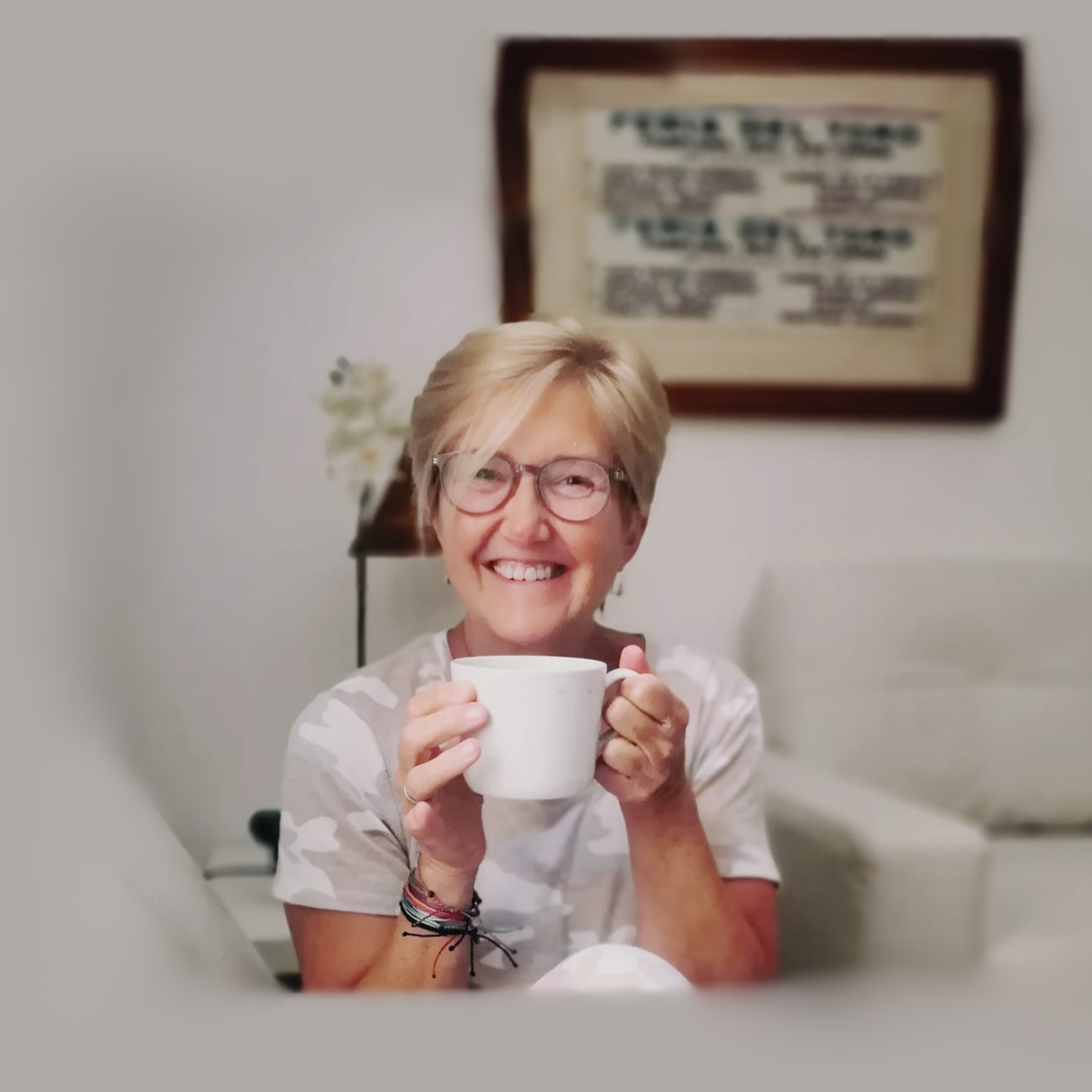 A smiling woman with short blonde hair and glasses holding a white mug, sitting in a cozy room with a framed picture on the wall behind her and a small table with a flower in a vase.