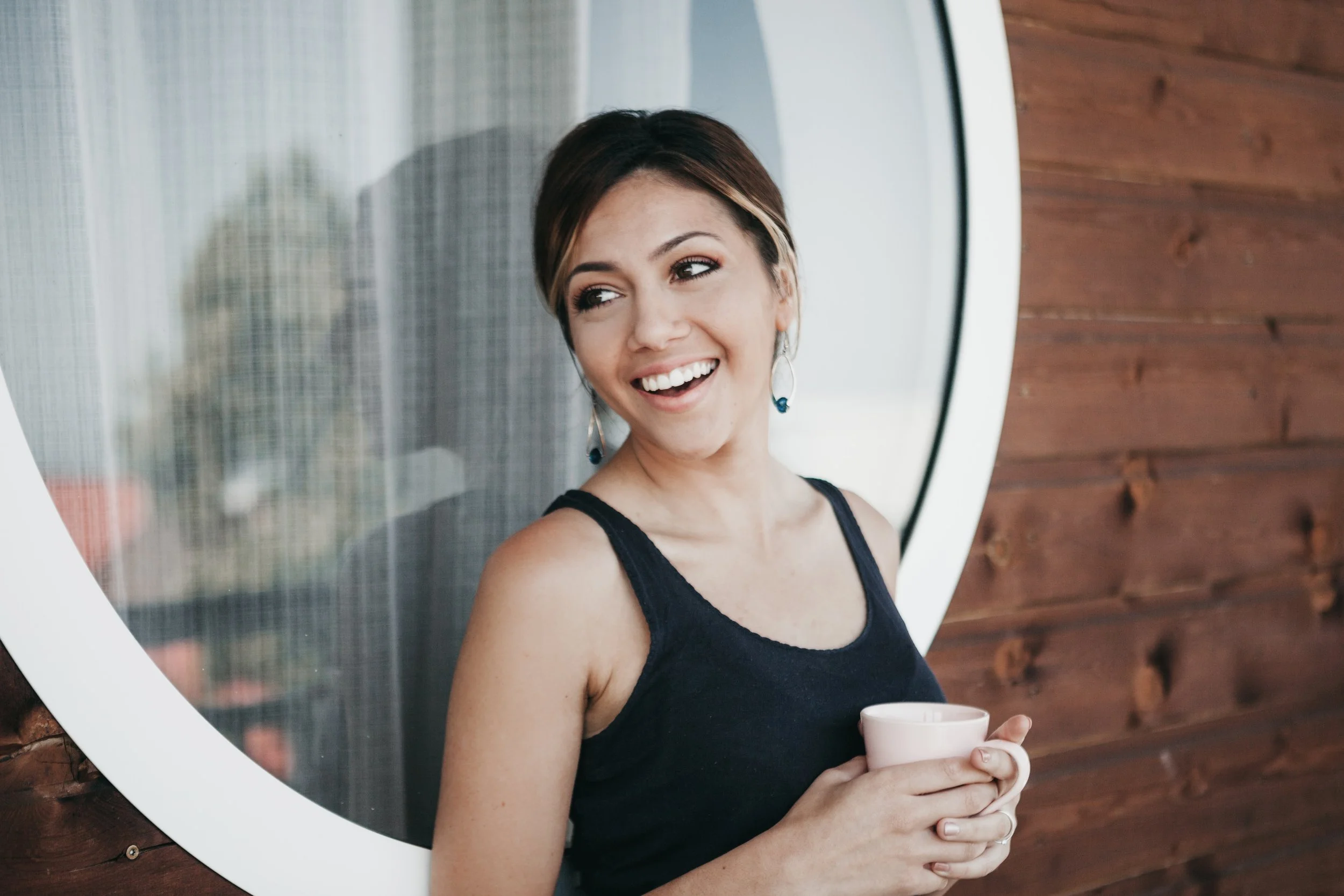 A woman with short hair and earrings smiling while holding a white mug, standing outside near a circular window and wooden wall.