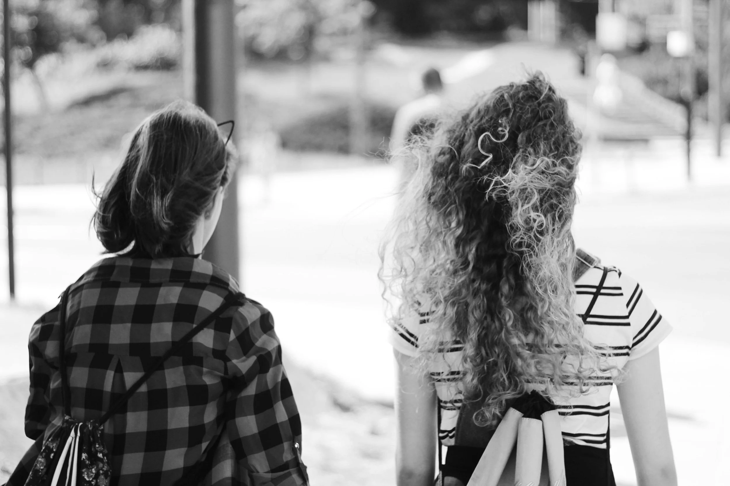 Two teens seen from behind standing outdoors, with one woman wearing glasses and a checkered shirt, and the other with long, curly hair wearing a striped T-shirt.