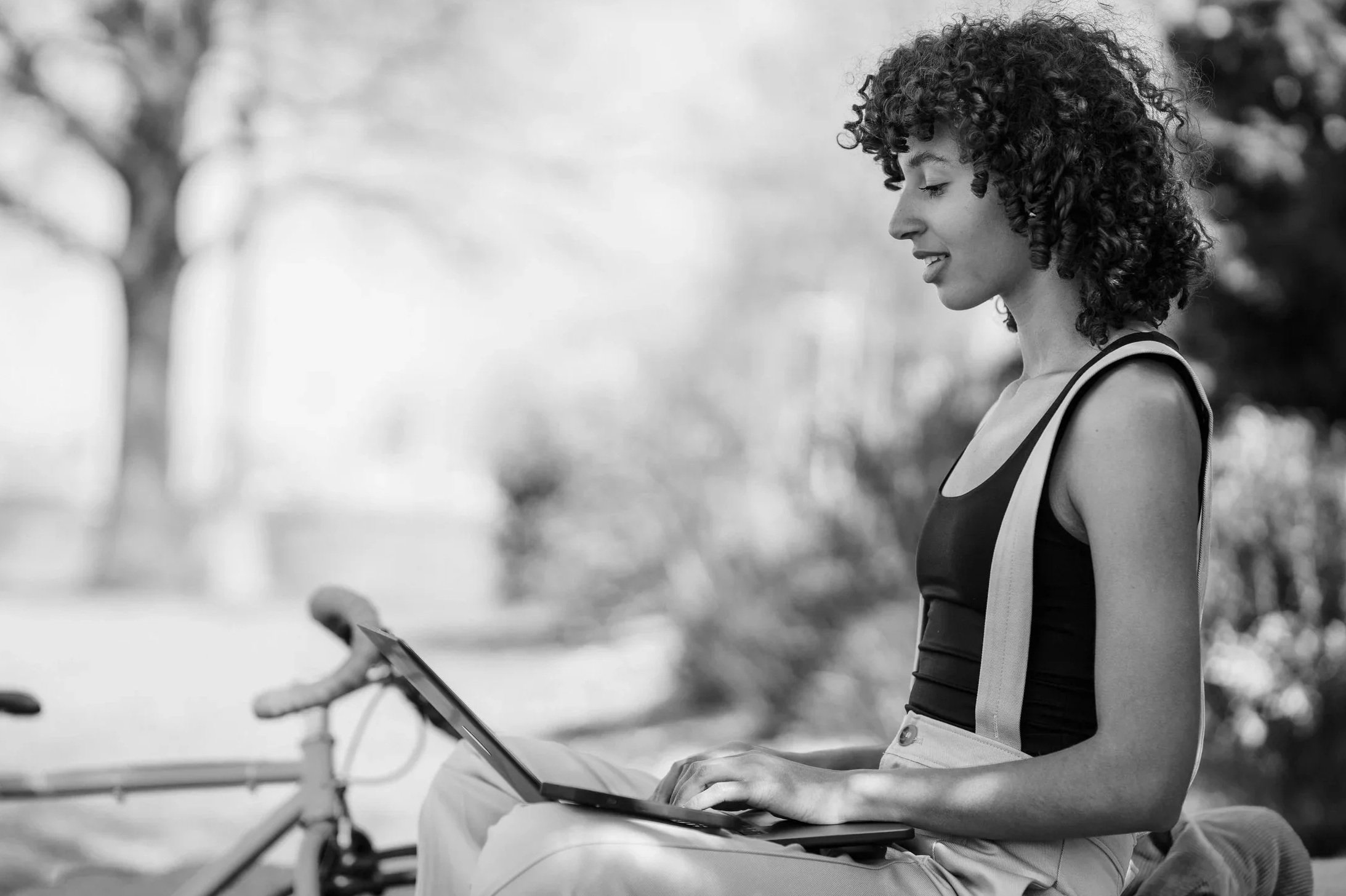 A young woman with curly hair sitting outdoors on a bicycle, using a tablet, smiling.