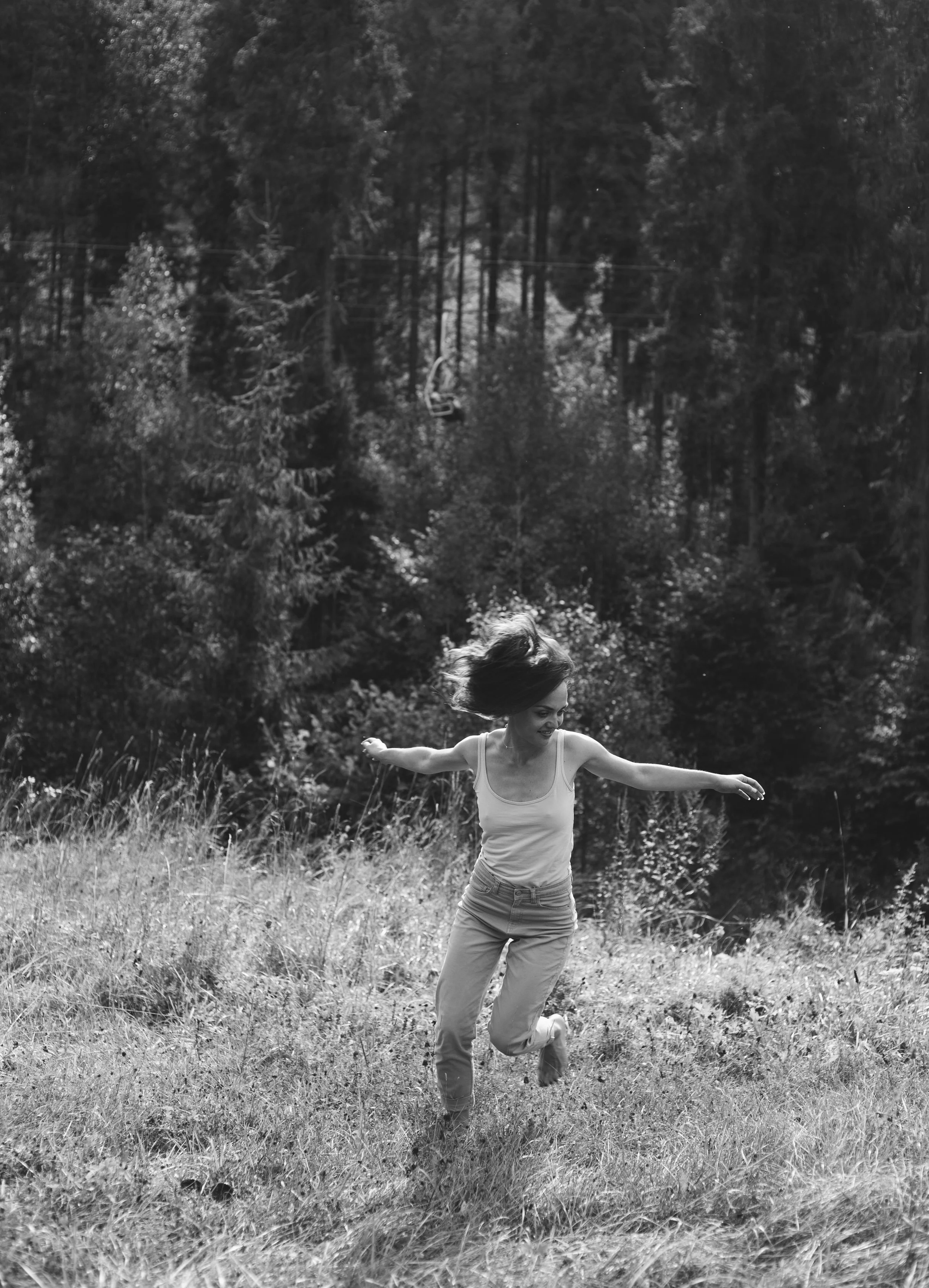 A woman with long hair dancing or jumping in a grassy field, with a backdrop of trees and a cable car in the distance.