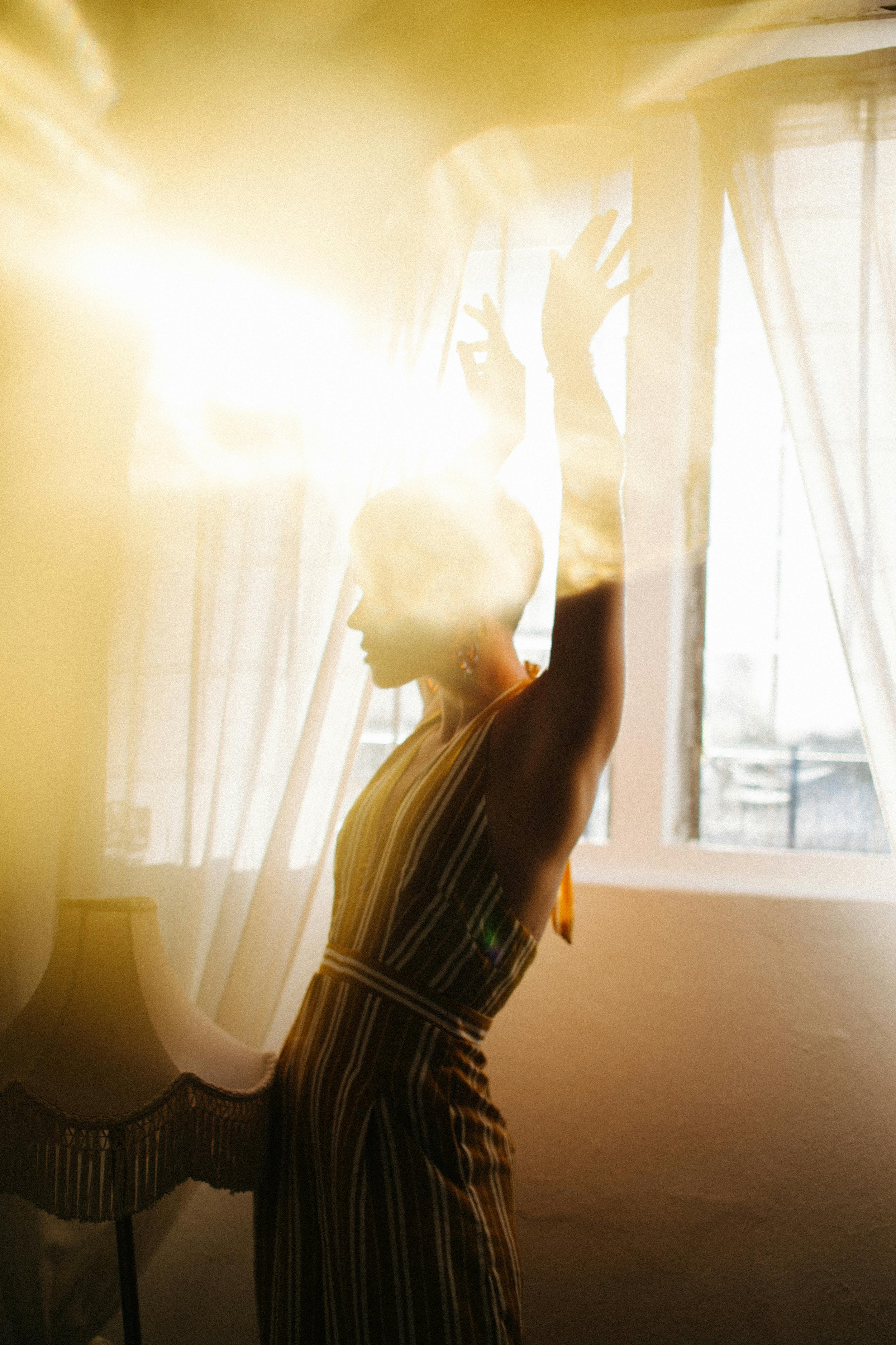 A woman standing indoors near a window with sunlight streaming in, raising her arms with hands forming a gesture, wearing a striped outfit.