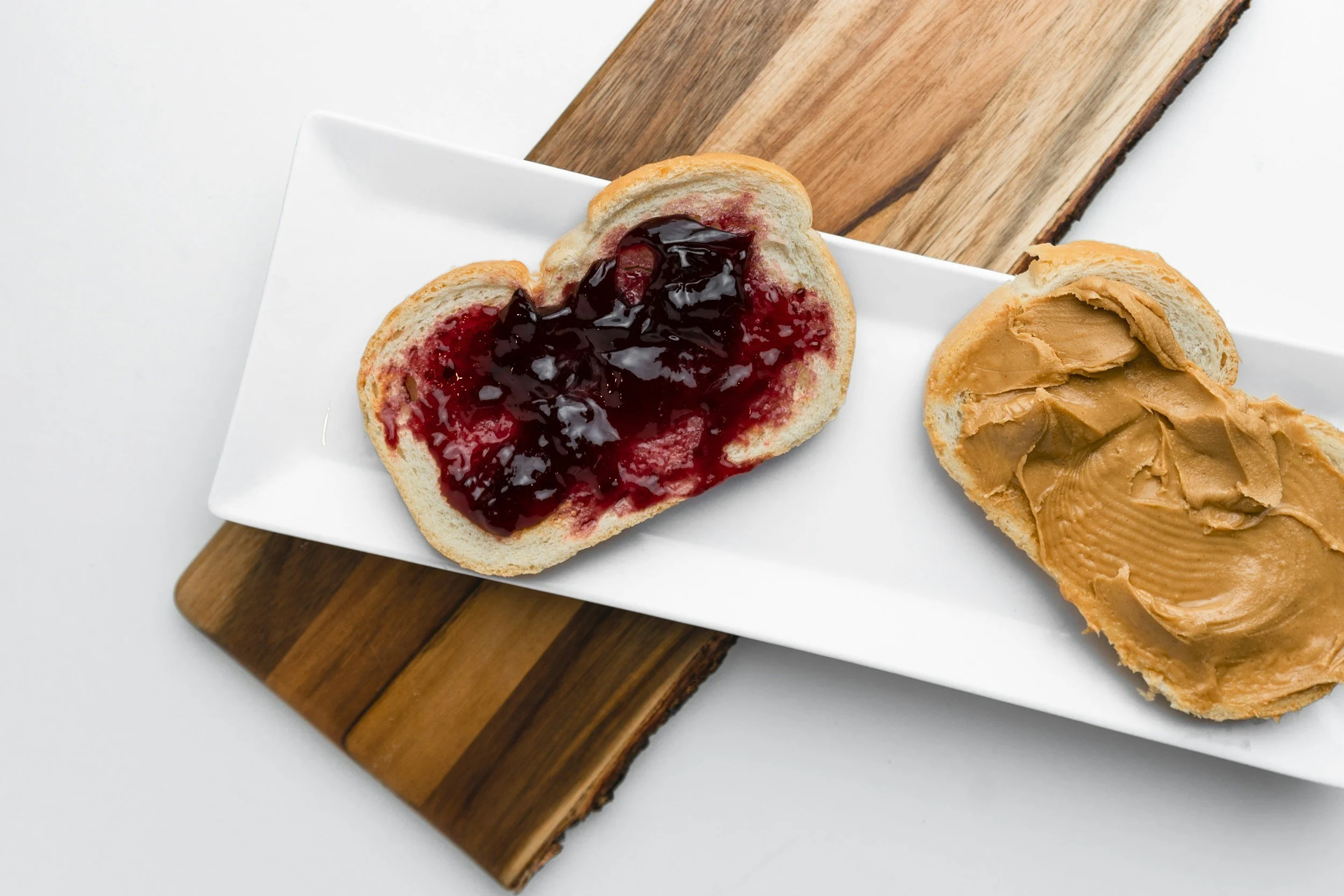 A slice of bread with jam and a piece of bread with peanut butter on a white rectangular plate, placed on a wooden board.