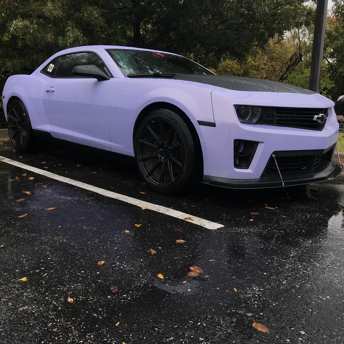 A white Chevrolet Camaro sports car with black wheels parked on a wet asphalt surface with fallen leaves, surrounded by trees with autumn foliage.
