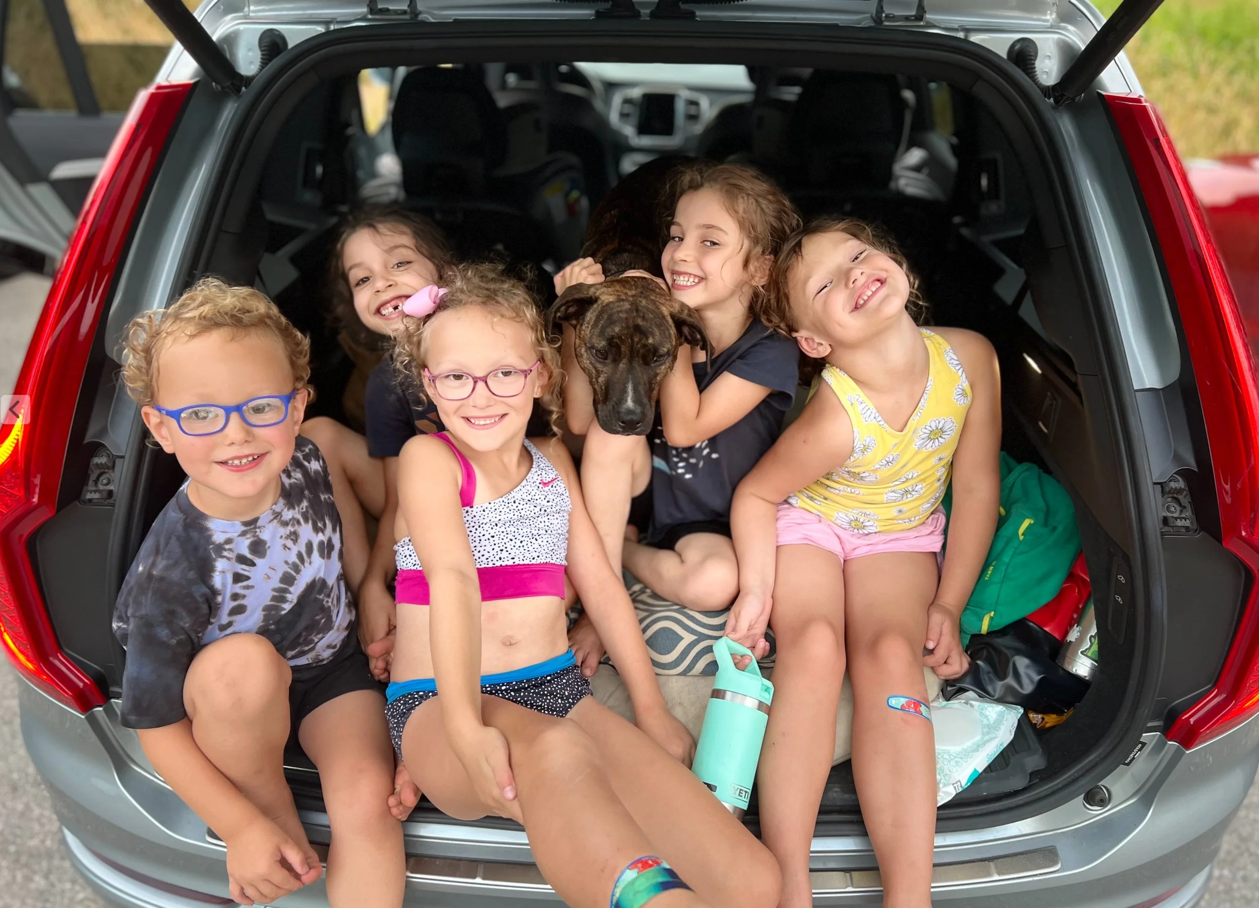 Six children and a dog in the open trunk of a car, smiling and posing for the camera during a trip or outing.