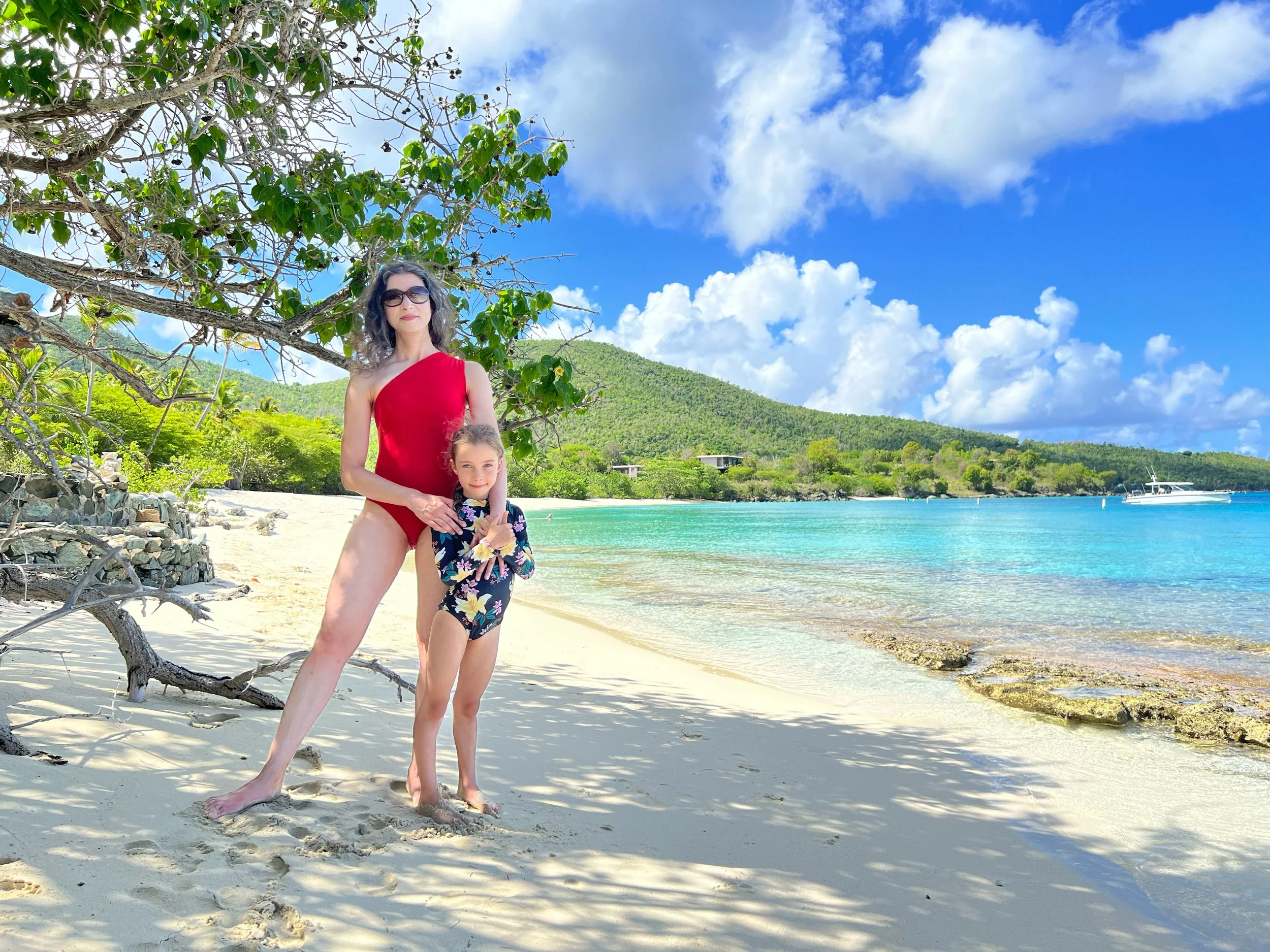 A woman and a young girl standing on a sandy beach with a tropical shoreline, green hills, and blue sky with clouds in the background