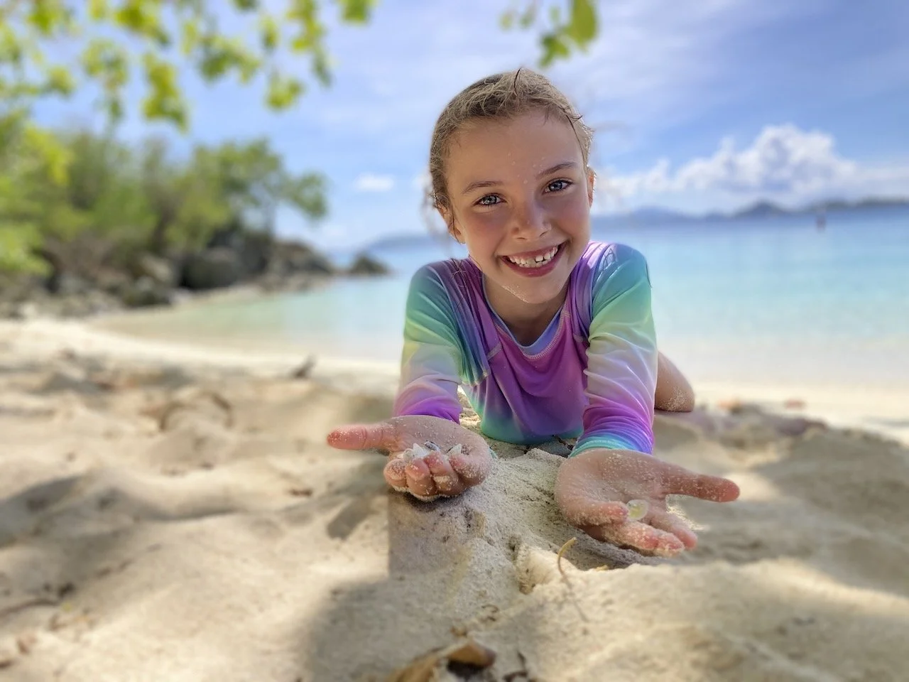A young girl lying on the beach in the USVI, smiling, wearing a rainbow-colored swimsuit, with the ocean, trees, and a cloudy sky in the background.