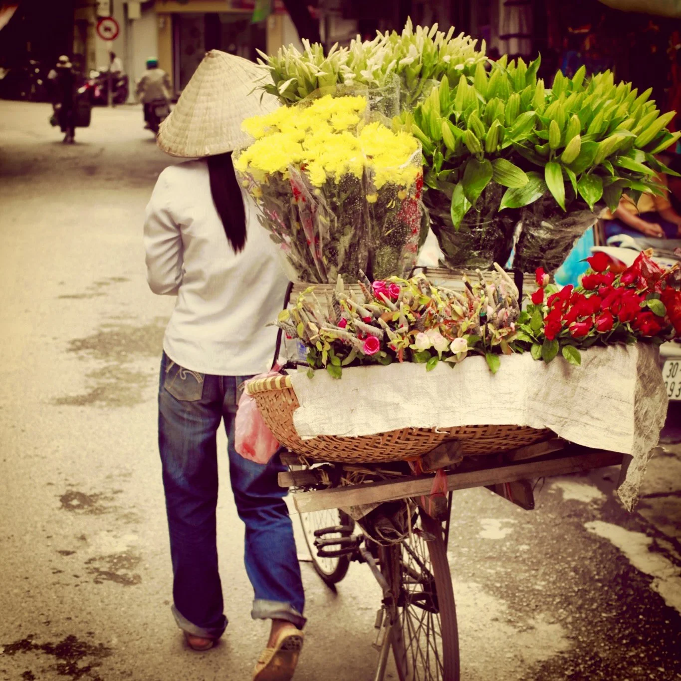 Person in a white shirt and jeans pushing a bicycle with a large basket of colorful flowers on the back on a street.