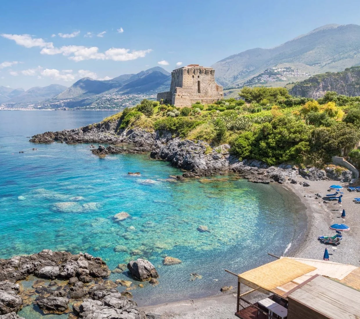 A scenic coastal view featuring a rocky shoreline, turquoise water, lush green vegetation, an ancient stone castle on the hill, mountains in the background, and a small beach with blue umbrellas and lounge chairs.