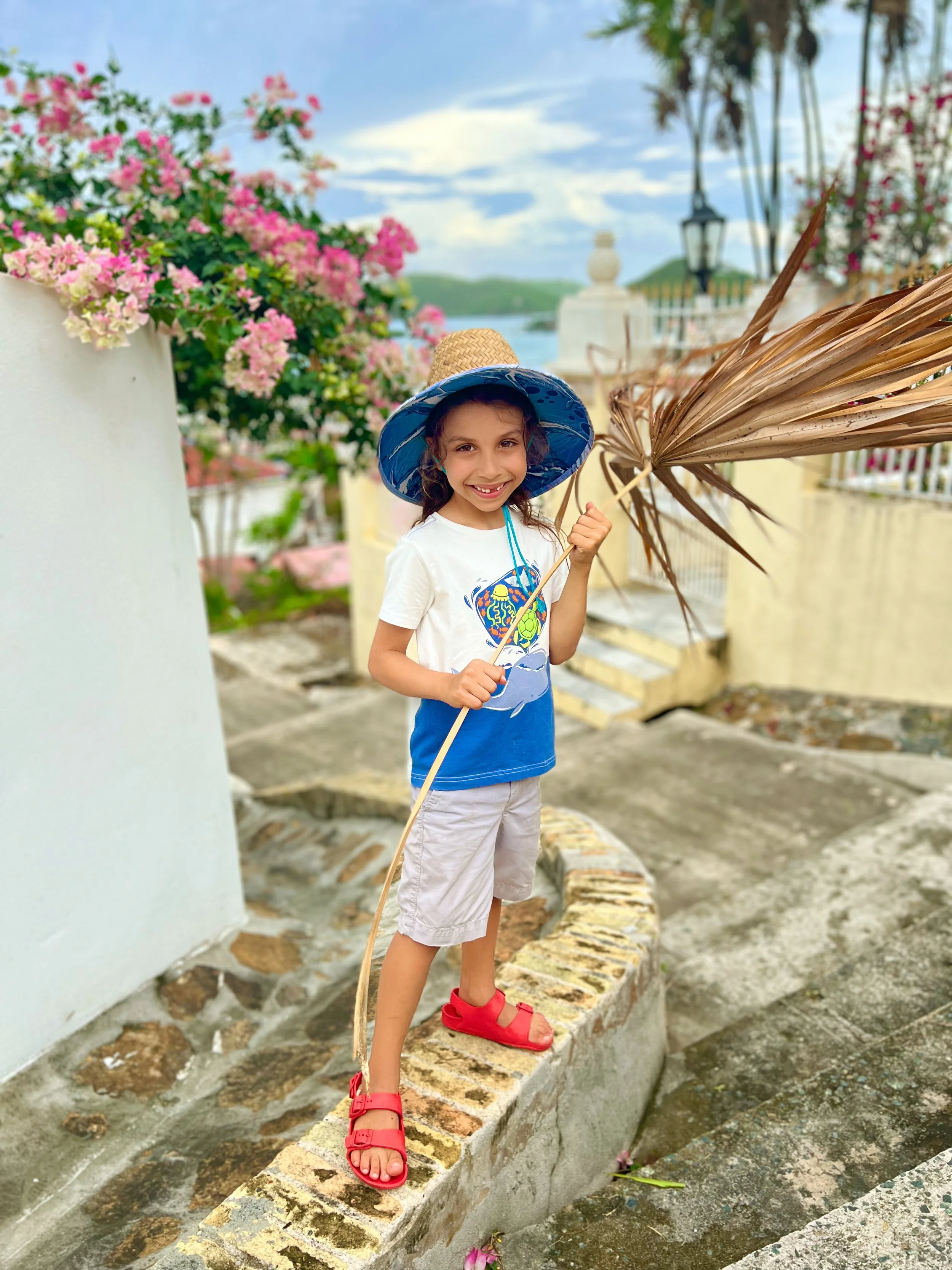 A young girl smiling while standing on a brick ledge outdoors, holding a dried palm leaf, wearing a blue sunhat, white and blue t-shirt, beige shorts, and red sandals. The background features pink flowers, trees, a streetlamp, and a cloudy sky over distant hills.