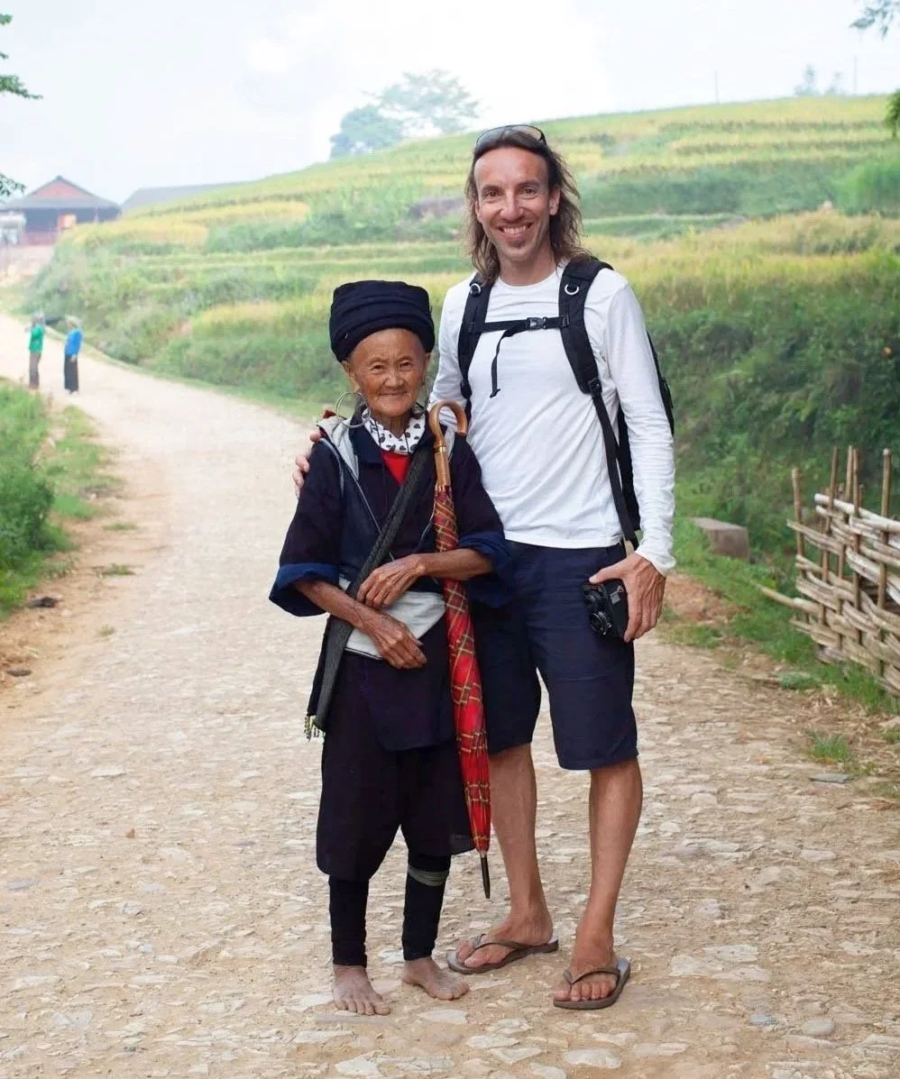 A young man and an elderly woman stand together on a dirt path in a lush, green rural area. The woman wears traditional clothing and holds an umbrella, while the man wears casual travel attire with a camera in hand. They smile at the camera with greenery and terraced fields in the background.