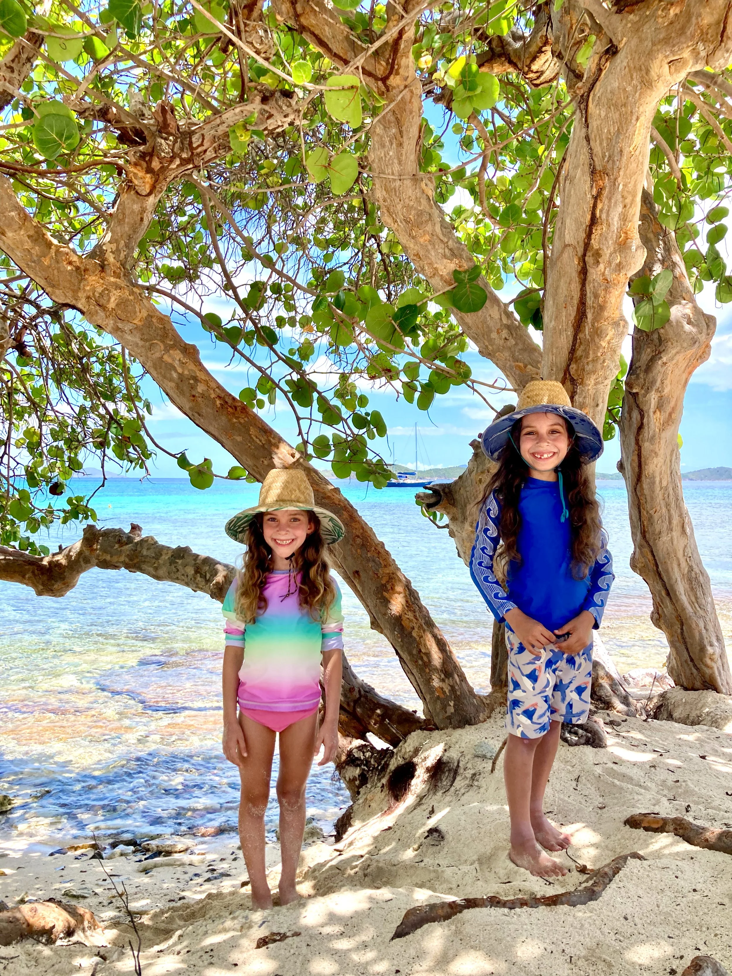 Two young girls with long, curly hair, wearing large sun hats and colorful beach clothing, standing on sandy beach near a tree with green leaves, with ocean water and sailboats in the background.