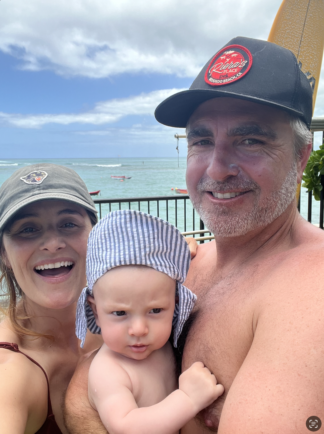 A happy family of three at the beach, with a woman, a shirtless man wearing a black cap, and a baby with a striped hat, standing in front of the ocean and boats, under cloudy sky.