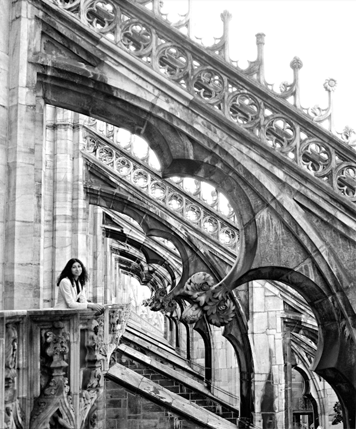 A woman sitting on a marble balcony of an ornate, Gothic-style building with intricate stonework and arched windows.