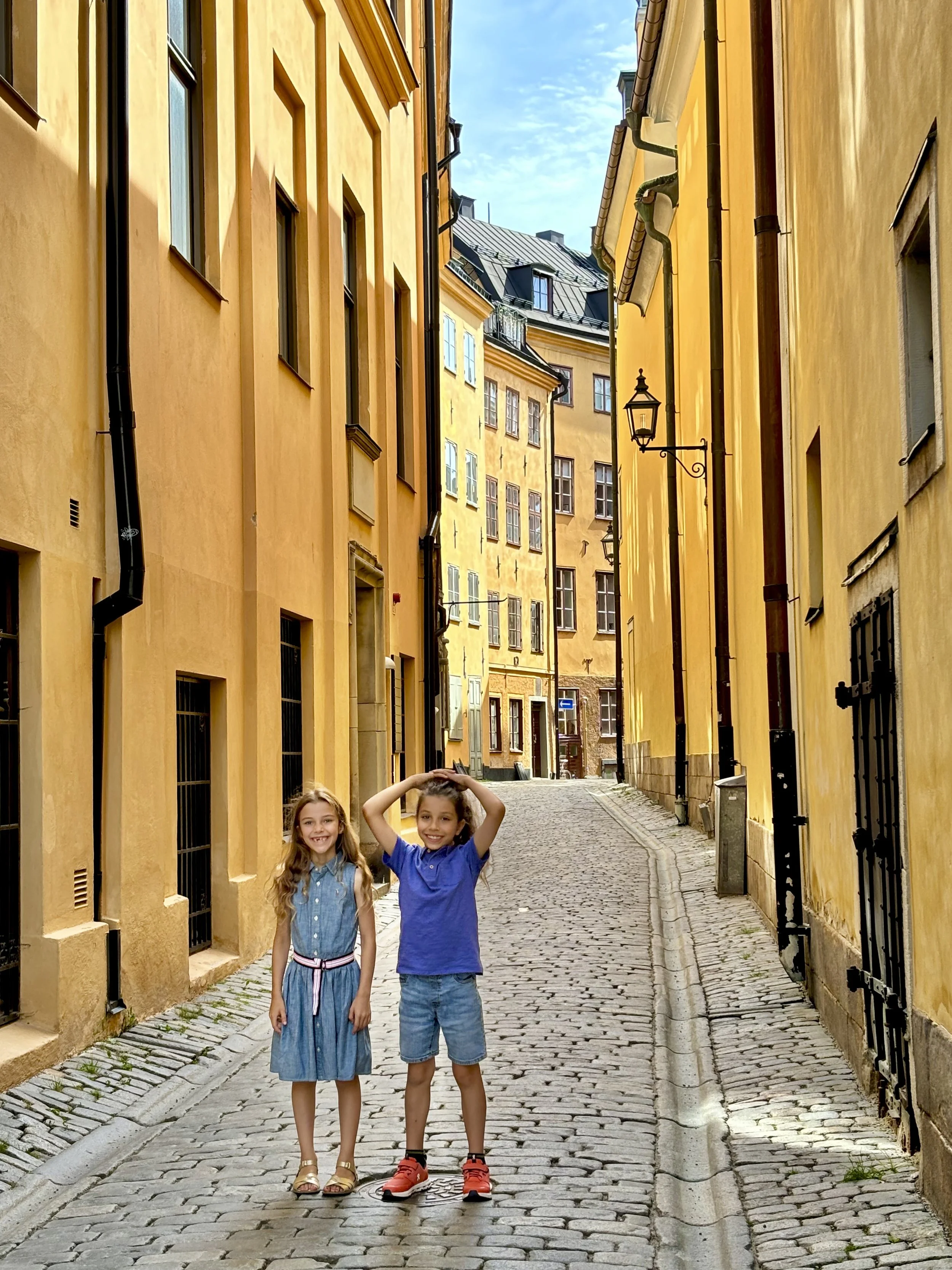 Two children, a girl and a boy, standing on a cobblestone street in Gamla Stan, Stockholm with yellow buildings on either side. The girl is wearing a blue dress and sandals, and the boy is wearing a blue t-shirt, denim shorts, and red sneakers.