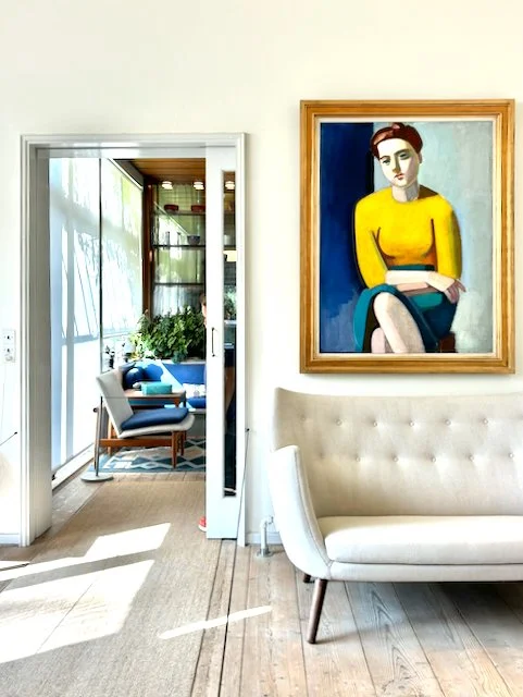 A modern living room with a white couch, a framed portrait of a woman in a yellow top hanging on the wall, and a doorway leading to a sunlit sitting area with blue chairs and large windows.