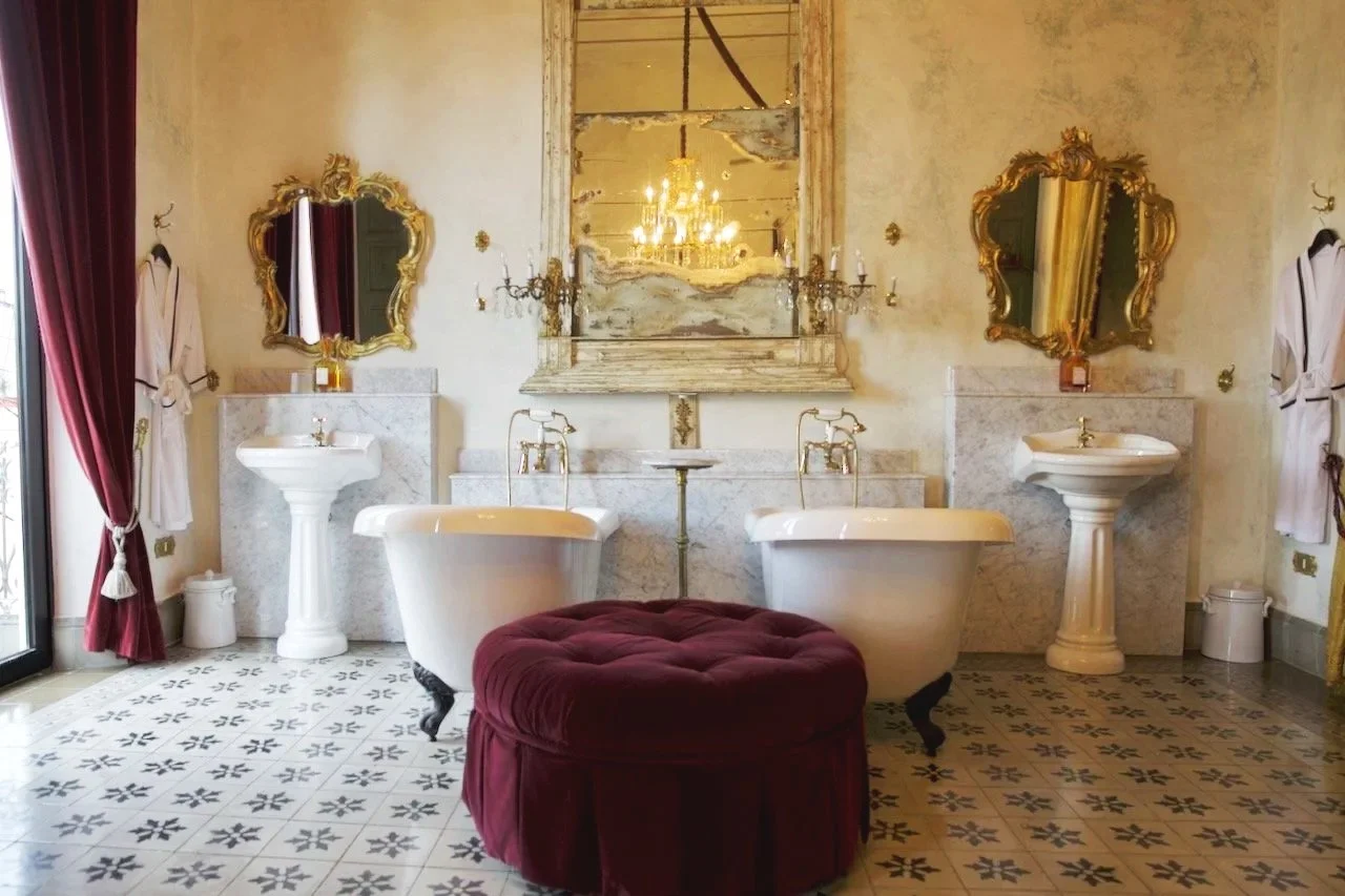 The elegant bathroom of Coqui Coqui Merida, with two pedestal sinks, a clawfoot bathtub, ornate mirrors, a chandelier, and vintage decor.