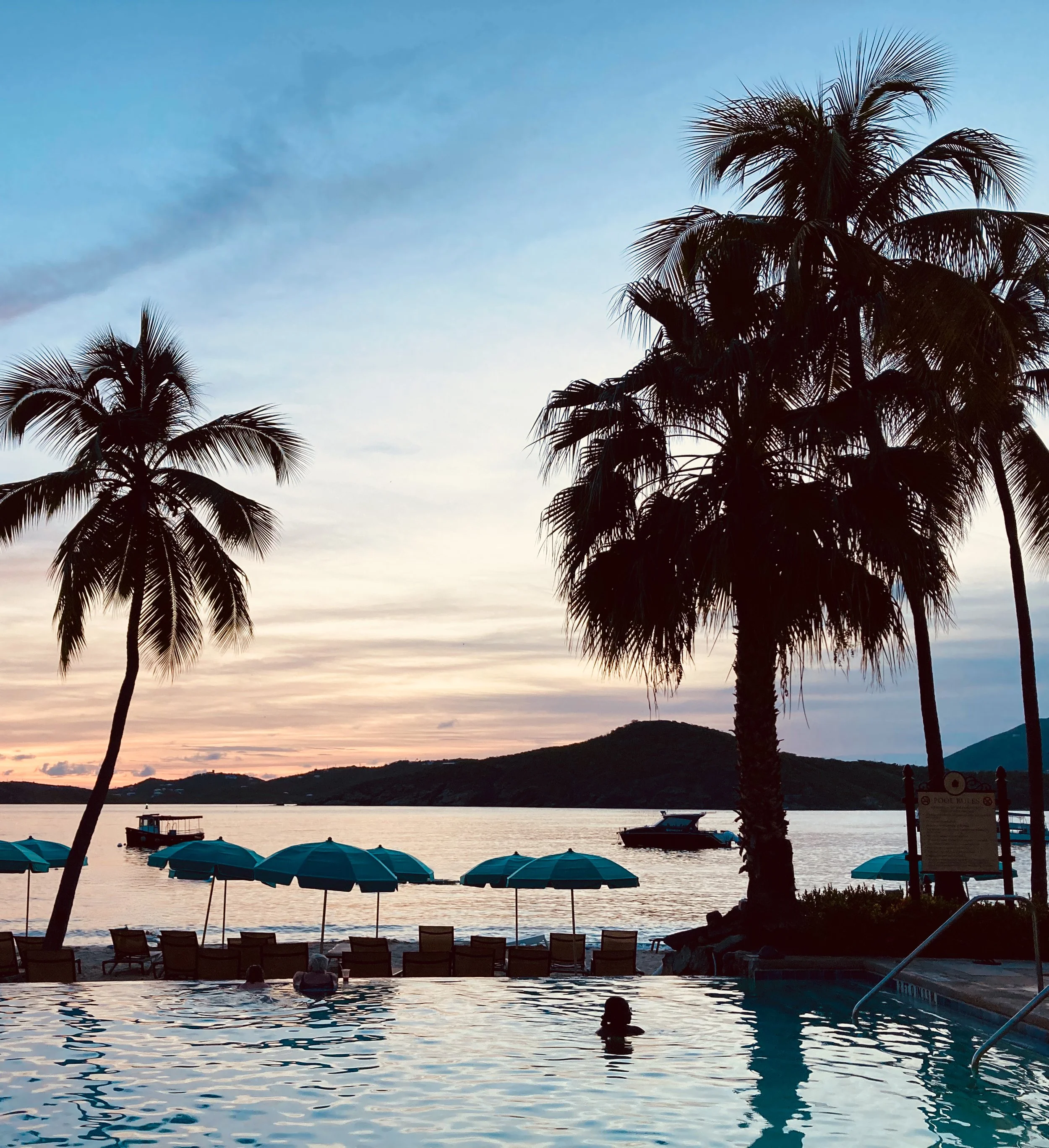 Sunset view over a beach with palm trees, lounge chairs and umbrellas near a swimming pool, with boats on the water in the background.
