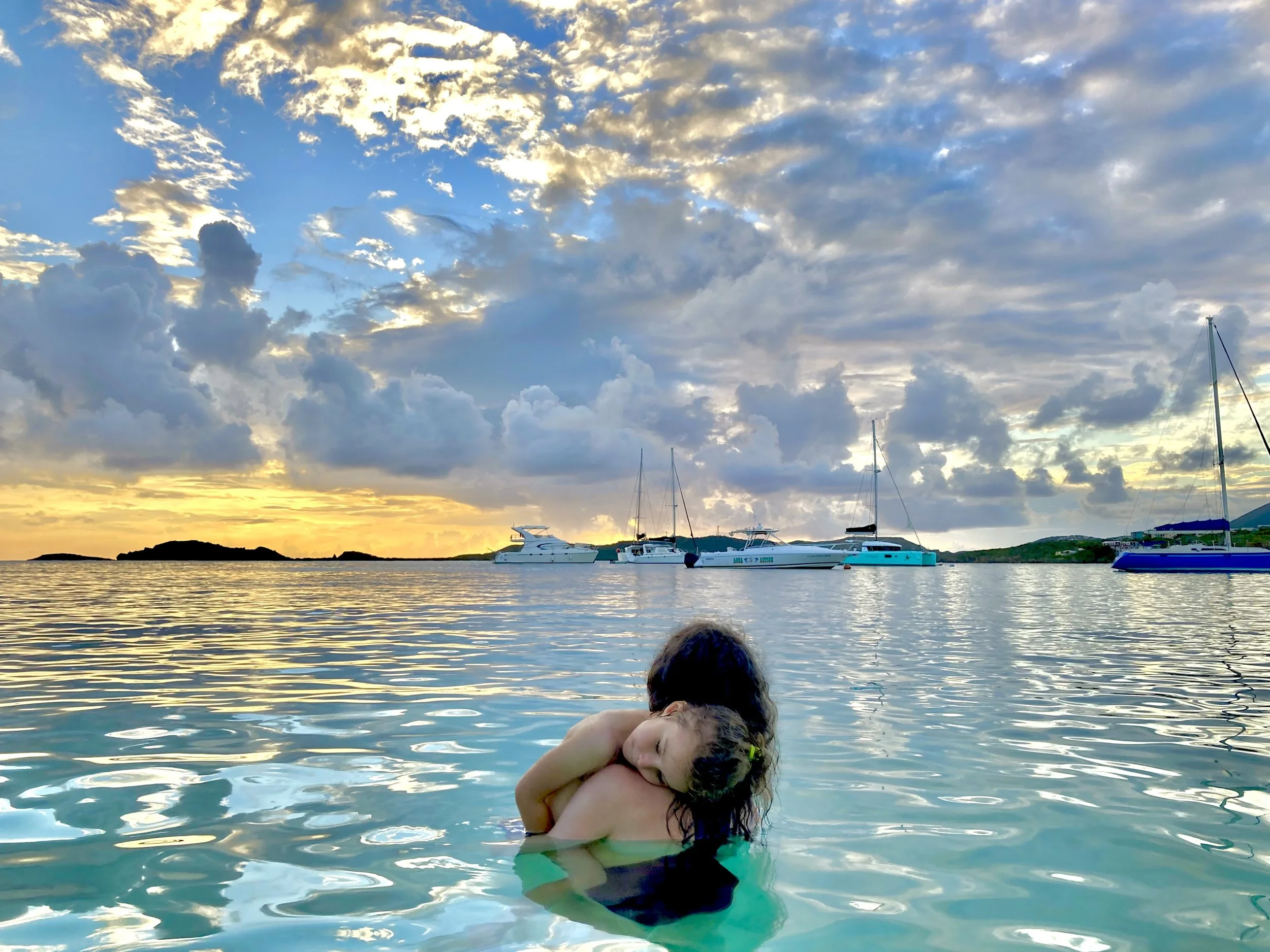 A woman and a child hugging in the water with boats and a sunset sky in the background.