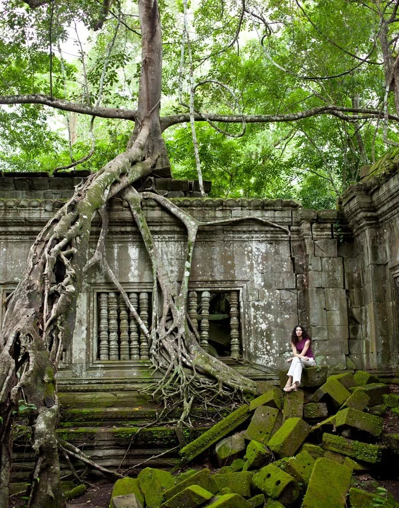A woman sitting on moss-covered stone blocks in front of an ancient temple wall entwined with large tree roots, surrounded by lush green foliage.