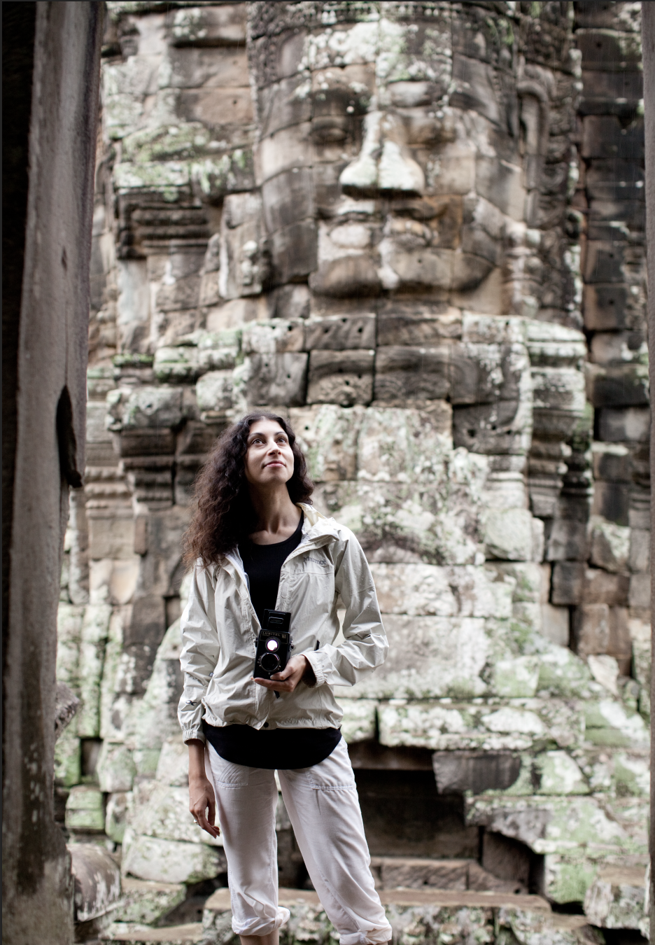 Woman with curly hair wearing a white jacket and holding a camera, standing in front of an ancient stone structure with a large carved face sculpture.