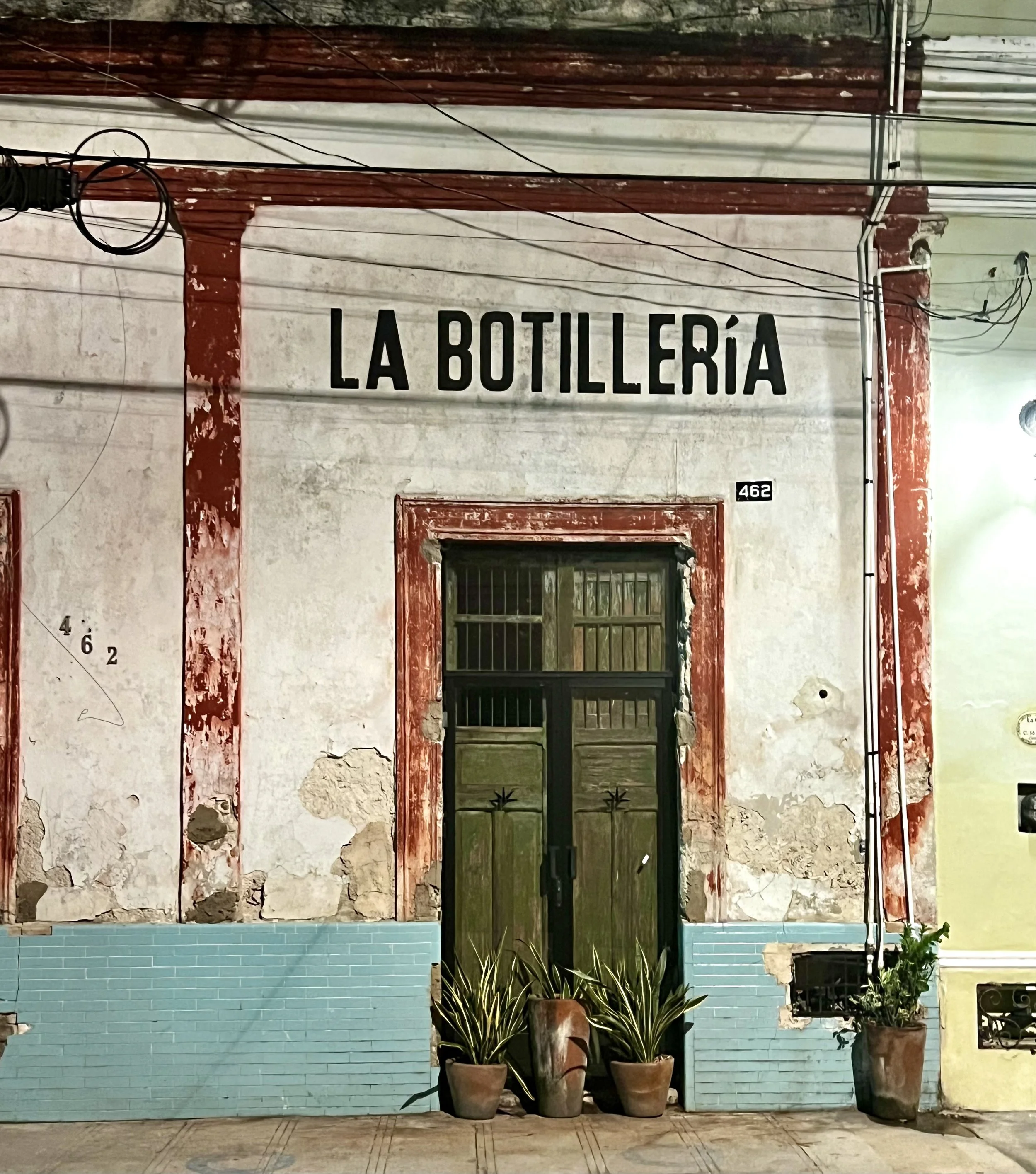 The weathered colonial facade of La Botillería at night, its painted plaster peeling around dark red terracotta trim, a dark green double door flanked by potted agave plants beneath bold black signage reading "La Botillería.