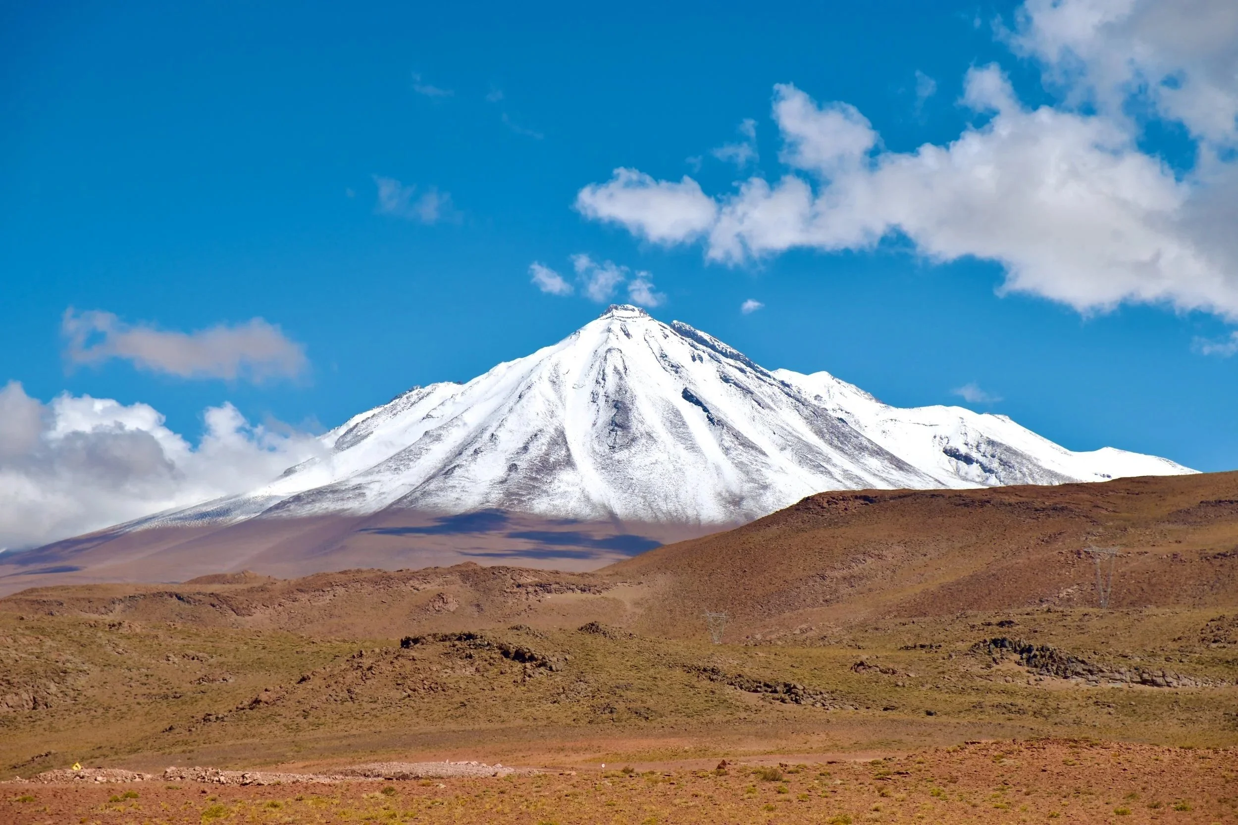 Snow-capped conical volcano rising above the rust-coloured Atacama altiplano under a deep blue sky, seen from the desert floor near San Pedro de Atacama.