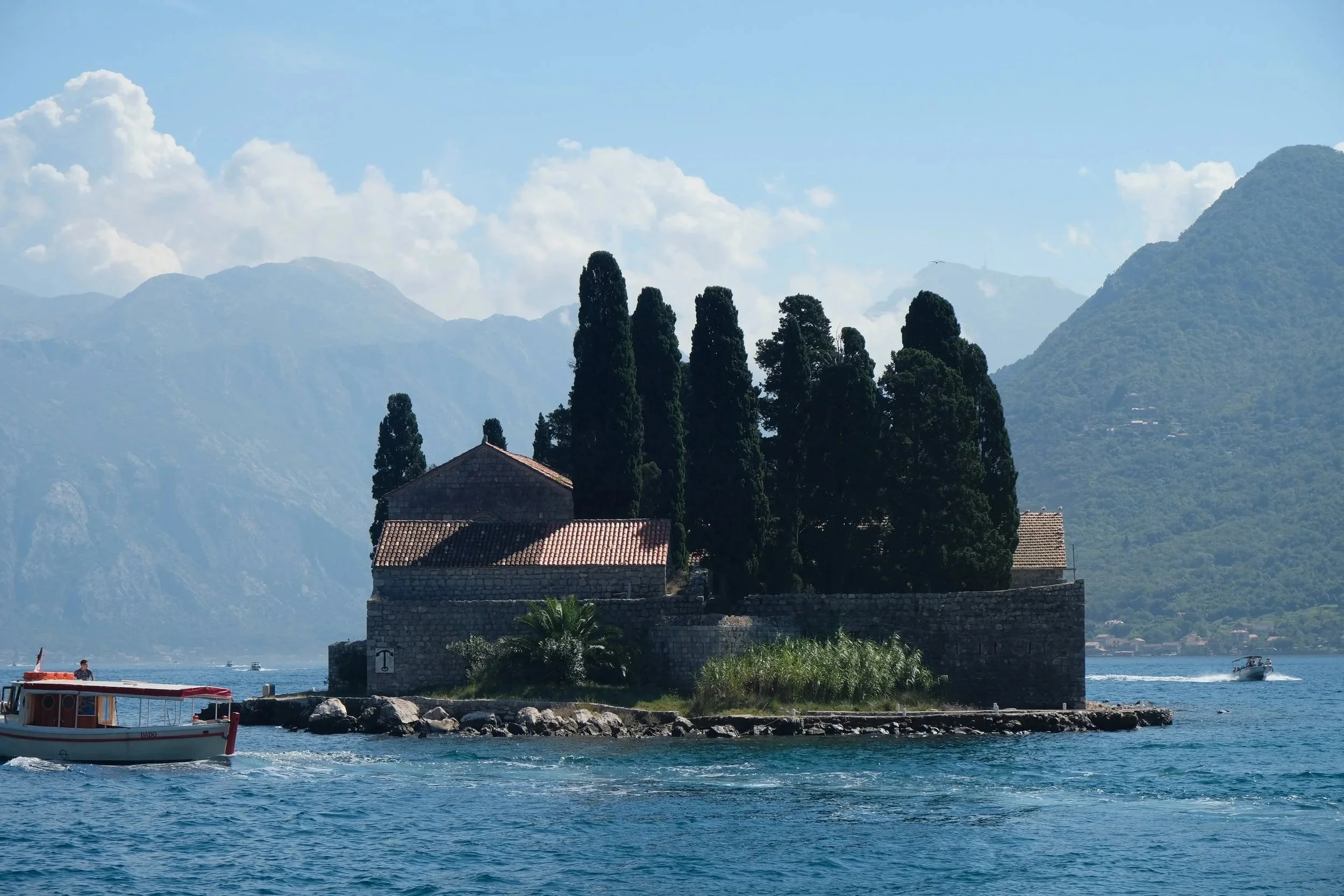 Small stone monastery on an island in a calm Adriatic bay with a wooden boat in the foreground and mountains behind — ALTEZA luxury cruise advisor