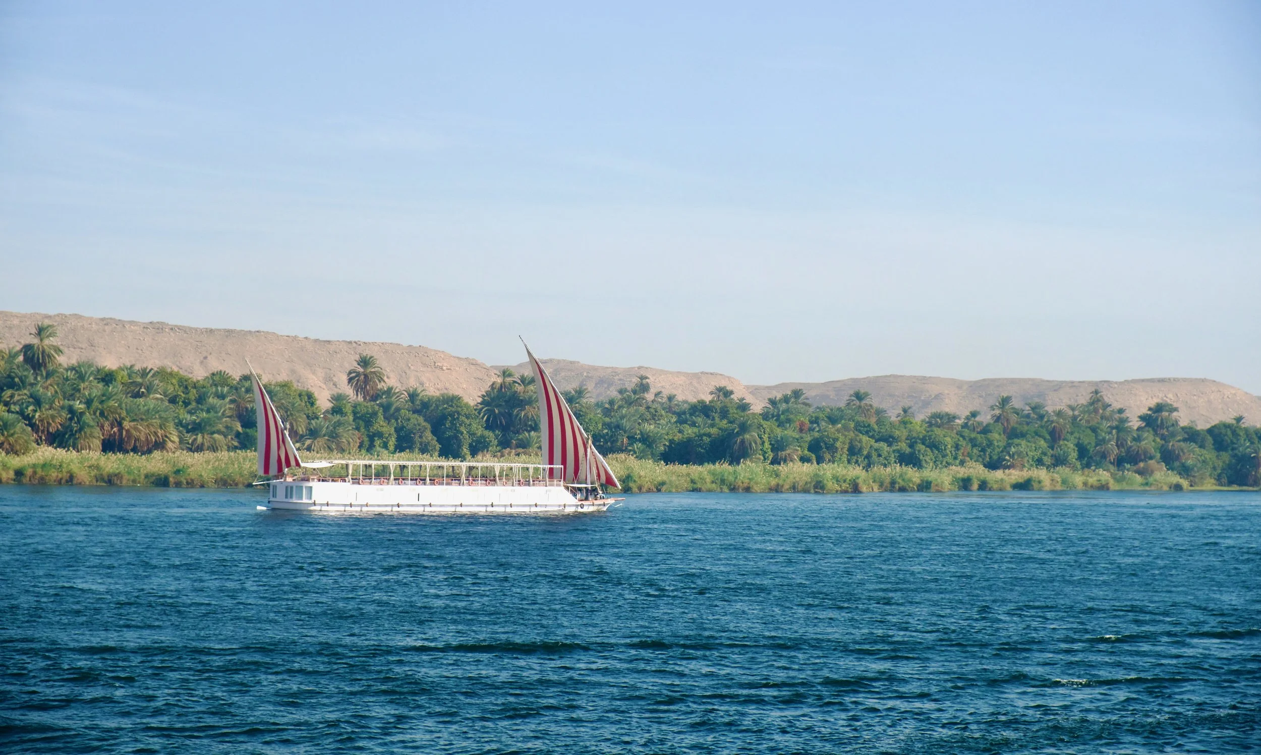 A traditional Egyptian dahabeya with red and white striped sails sailing the Nile, with a lush green riverbank, palm trees, and sandstone cliffs stretching behind it.