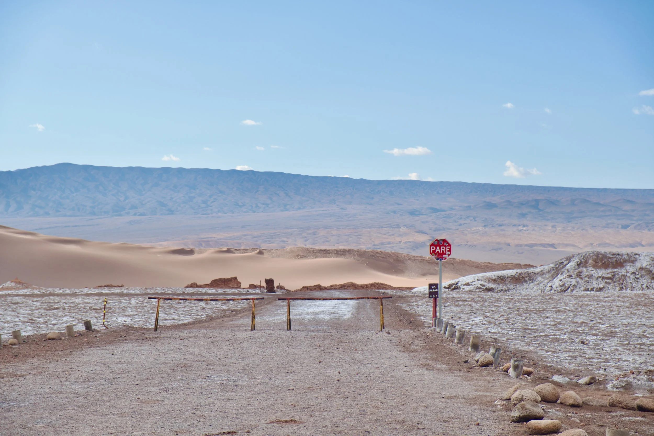 A gravel desert road leads to a metal barrier bearing a red PARE sign, with sand dunes and a distant mountain range stretching beyond into the Atacama Desert.