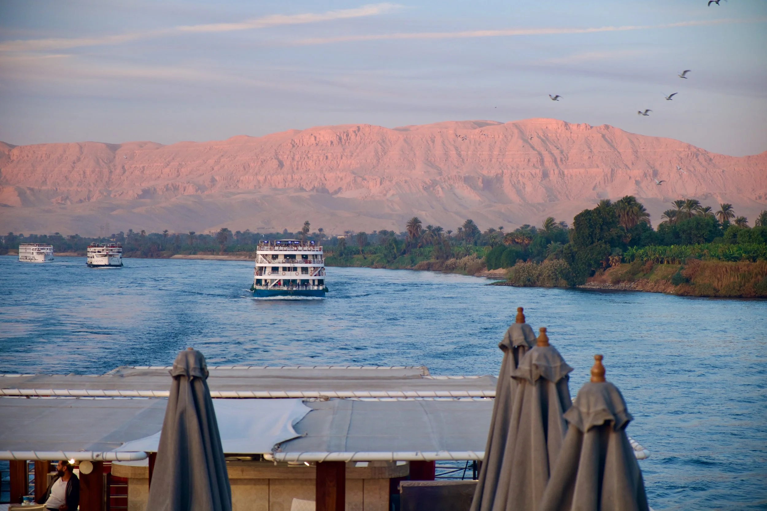 View from a Nile cruise ship's deck at dusk, with closed parasols in the foreground, two cruise vessels on the river mid-distance, and rose-lit desert cliffs catching the last of the day's light.