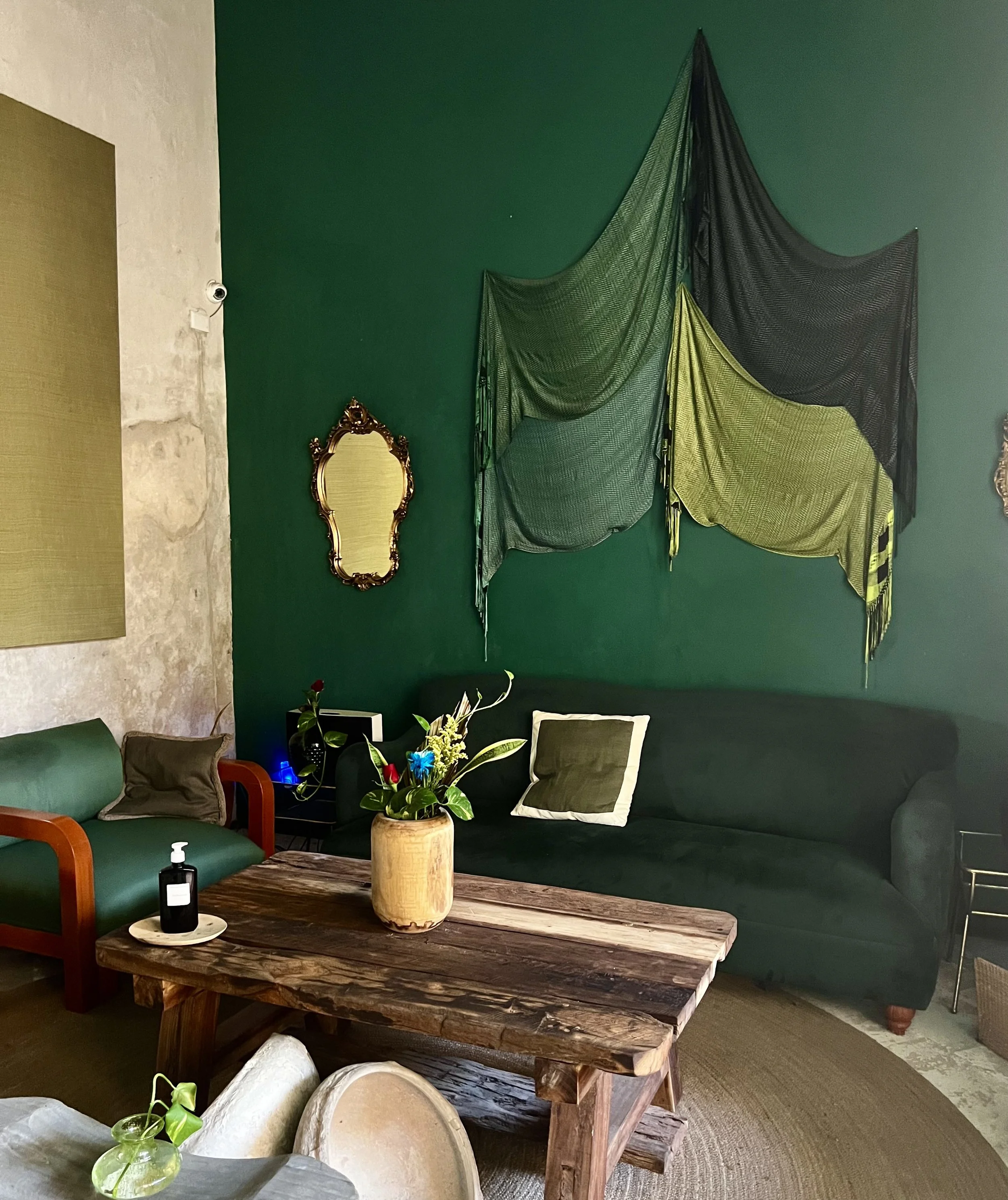 Interior of Ya'ax Hotel Boutique in Mérida, featuring a deep forest green painted wall with draped textile installation, a gilt baroque mirror, a dark green sofa, and a heavy rustic wooden coffee table on a woven rug.