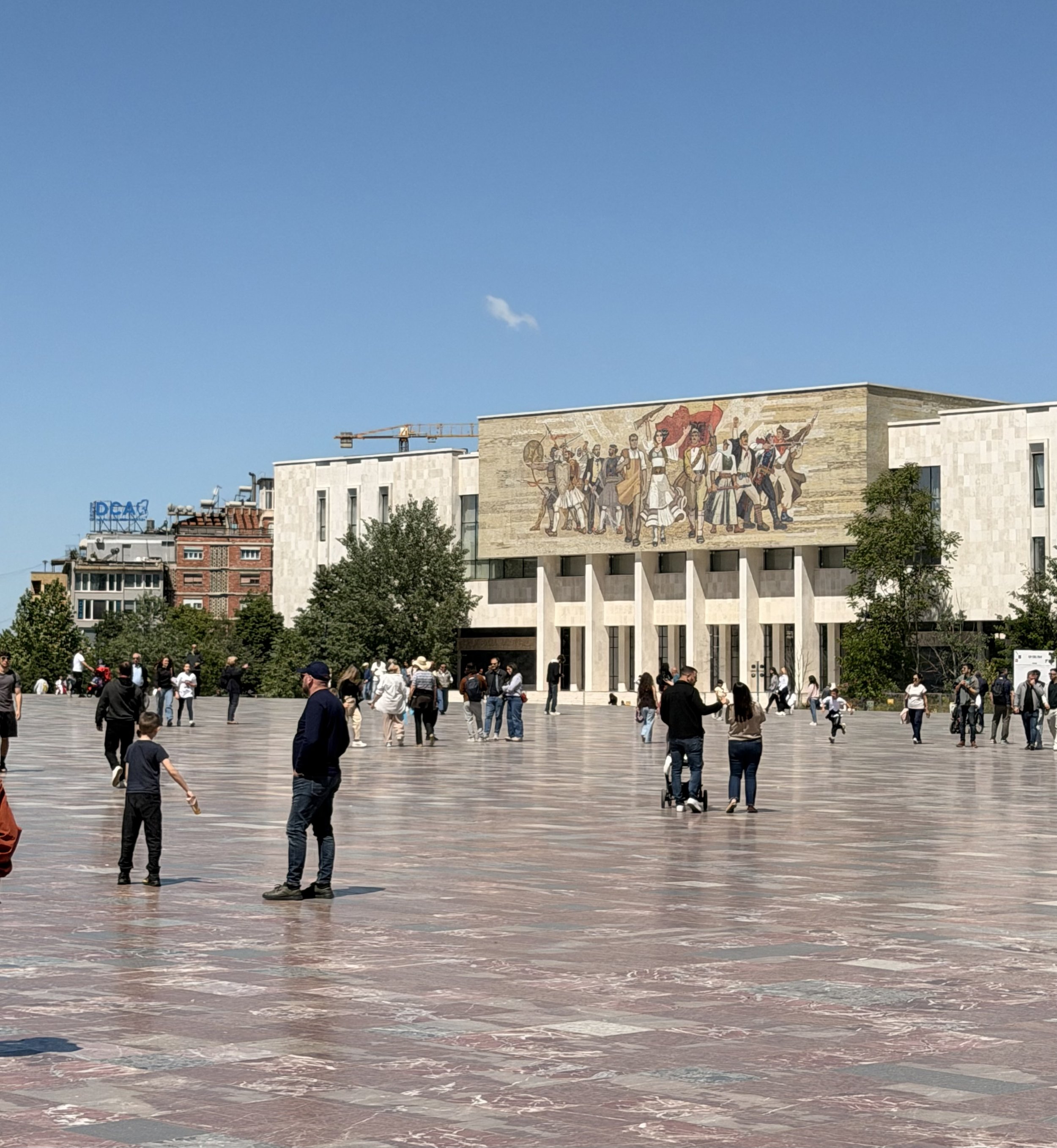 Visitors cross the wide marble expanse of Skanderbeg Square in Tirana on a clear day, with the National History Museum's facade and its large figurative mosaic mural visible in the background.