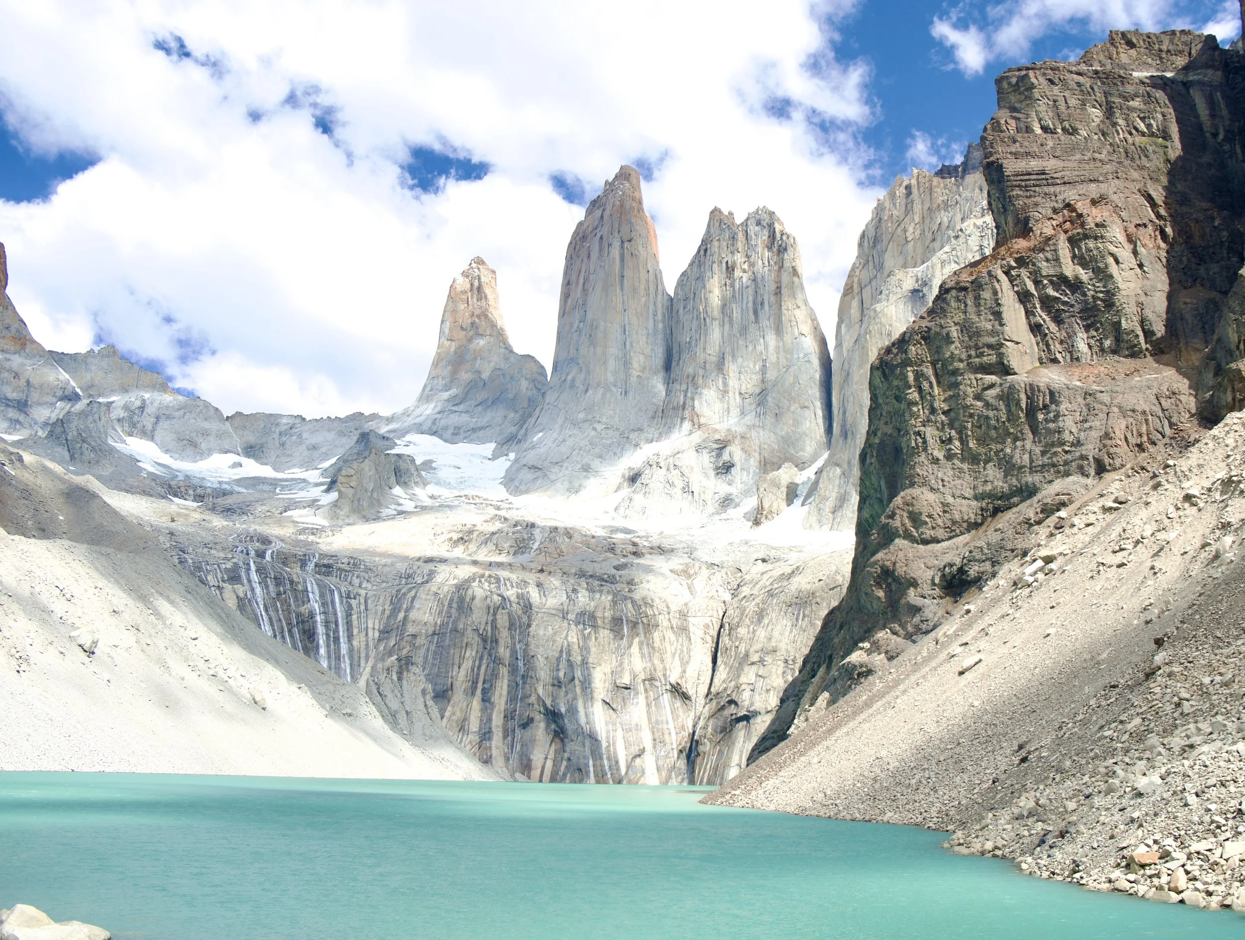 The three granite towers of Torres del Paine rising steeply above a turquoise glacial lake, with snowfields, cascading meltwater, and cloud-wrapped peaks behind them in Chilean Patagonia.