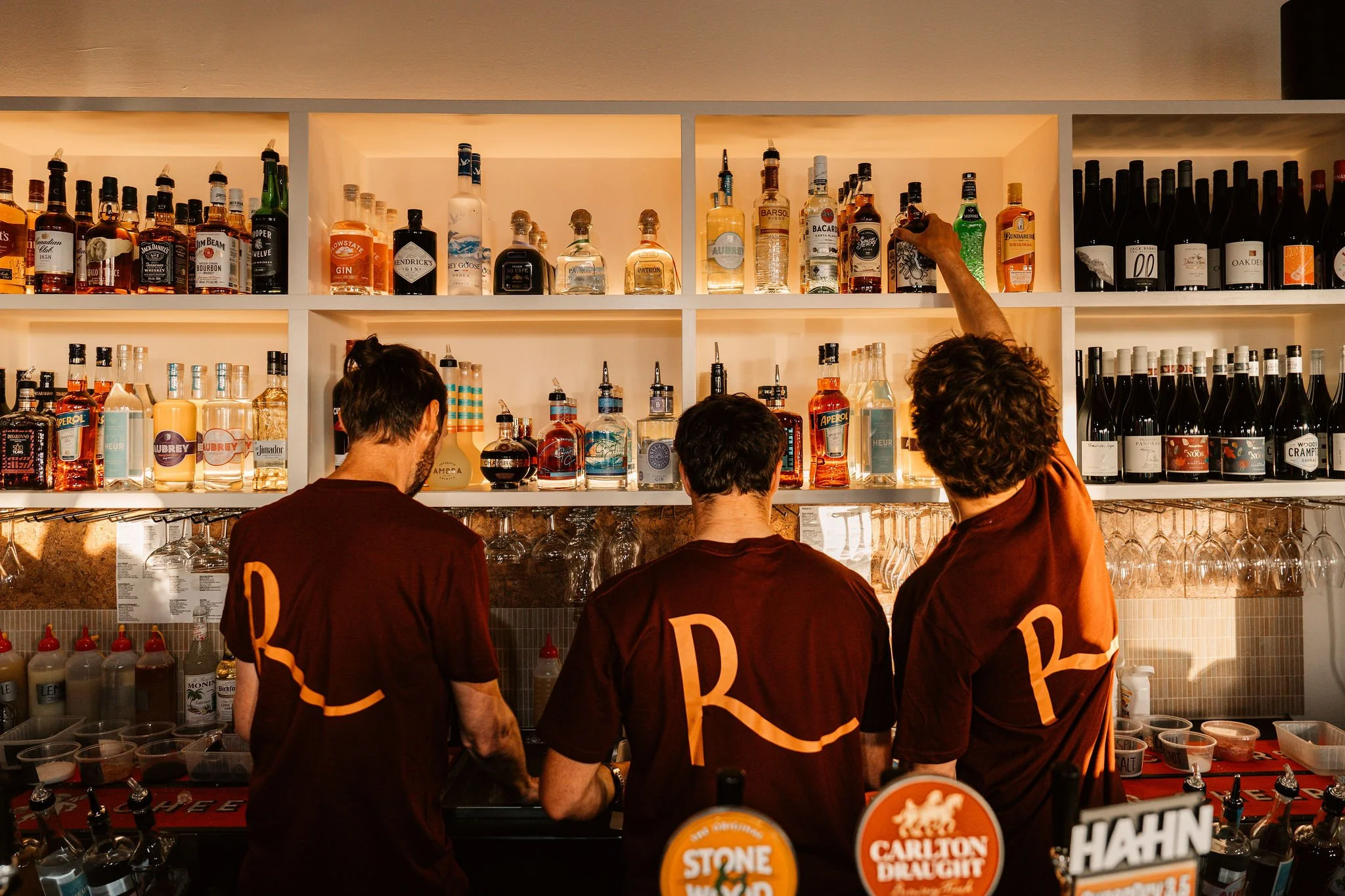 Three bartenders in burgundy uniforms with a large 'R' logo on the back on their top. They are working in a bar, reaching for spirit bottles on a shelf