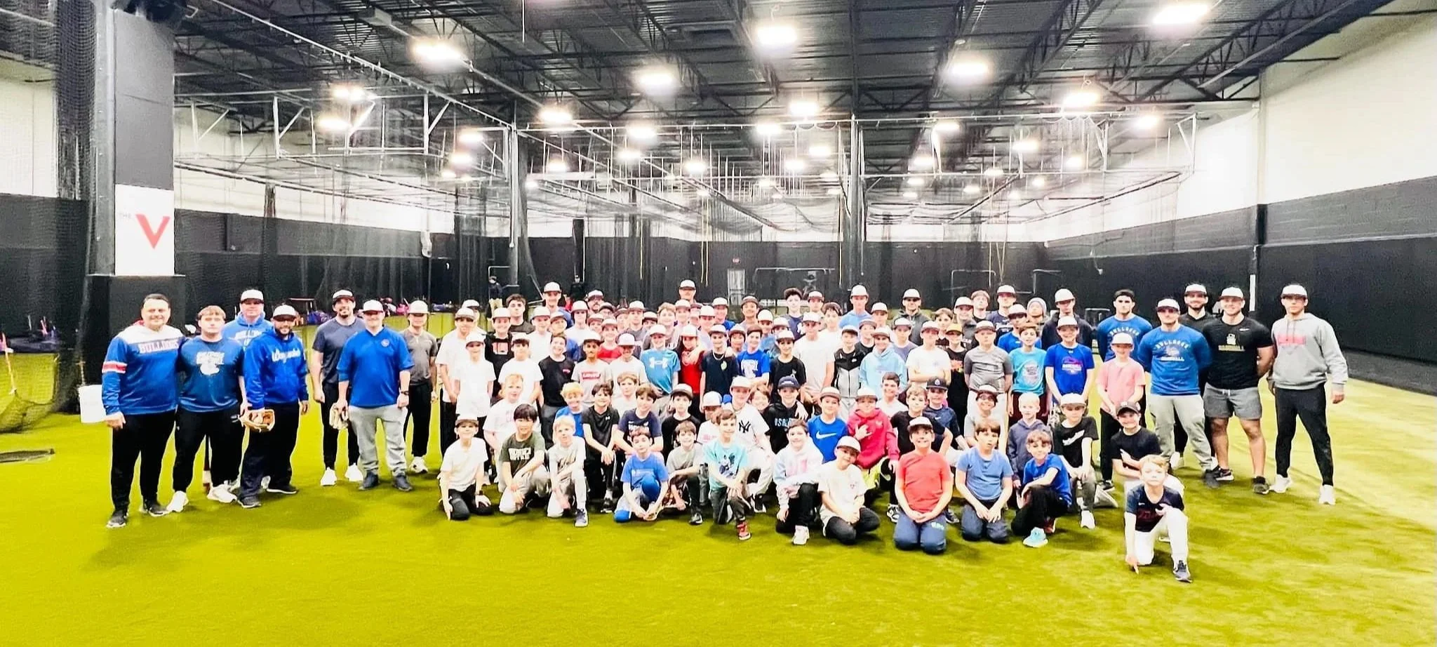 A large group of children and adults in an indoor sports facility, wearing baseball caps and athletic clothing, posing for a group photo on a green turf field. Vector Sports & Performance. Bulldogs Next Gen.