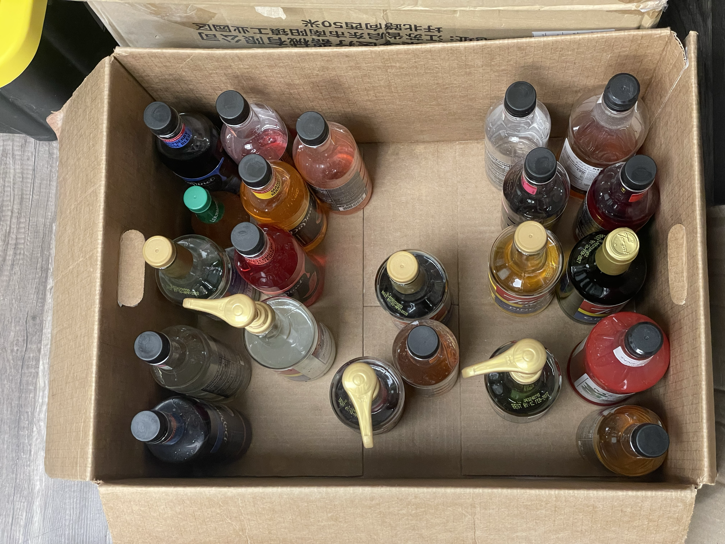 A cardboard box filled with various bottled beverages, some with spray nozzles, placed on a wooden surface.