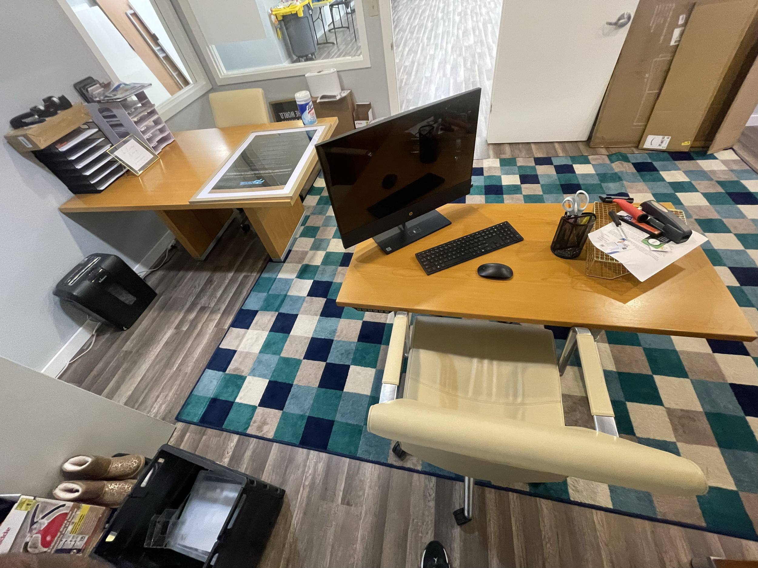 Office reception area with wooden desks, a computer monitor, keyboard, mouse, and office supplies. There is a colorful checkered rug, a chair, and a door leading to another room. Various office items include scissors, a highlighter, and documents. A 
