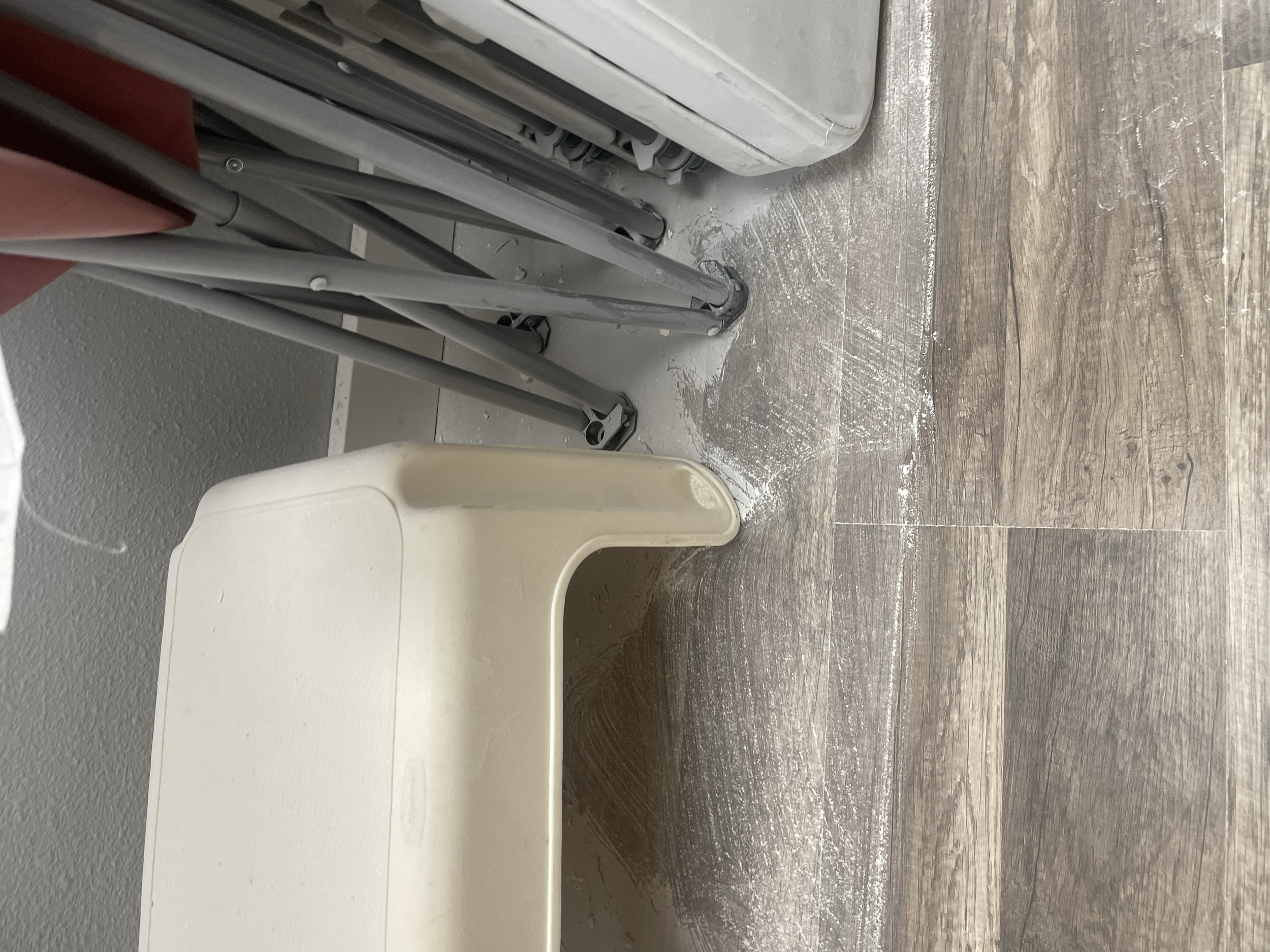 A corner of a room showing folded metal chairs, a beige plastic table, and wooden flooring with some white paint on it.